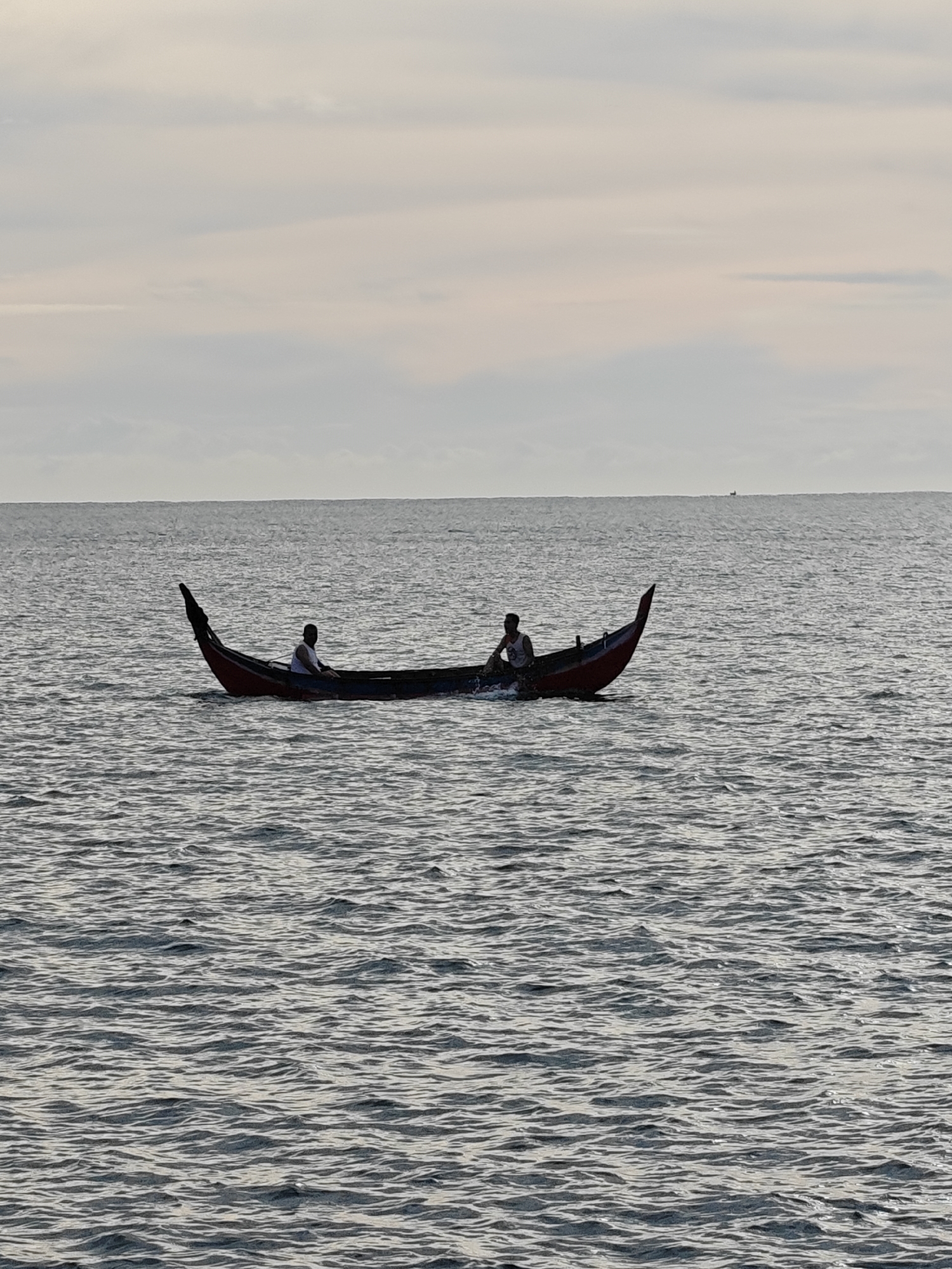 Two people are in a red boat on the sea. The sky is overcast. The location is Lhokseumawe, Indonesia.