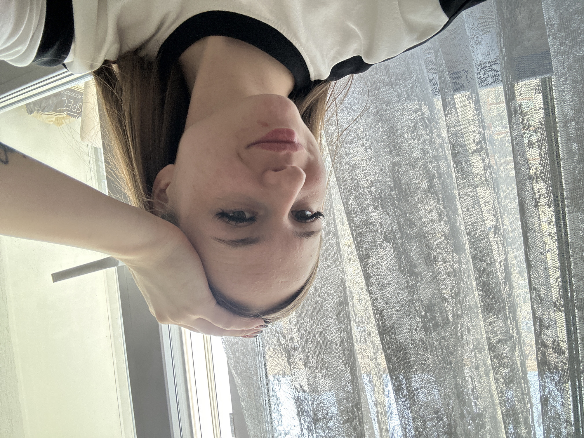 The image is a portrait of a young woman, captured from a high angle and presented rotated 90 degrees counter-clockwise, making her appear inverted. She has long, straight, light brown hair and fair skin, and is wearing a black and white long-sleeved top. Her head is tilted, and she rests her chin and the side of her face on her hand. Her eyes are open, looking towards the top of the frame (when viewed with correct orientation), and her lips are slightly parted, adorned with a hint of lip gloss. Her expression is neutral, possibly pensive or thoughtful.
The setting is indoors, with bright daylight entering through a window covered by sheer, light-colored curtains. These curtains feature a distinctive lace-like or honeycomb texture. A blurred outdoor scene is visible through the curtains and window, suggesting urban or residential surroundings.
A notable detail is visible through the window on the left side of the frame (which would be the lower left if the image were properly oriented). Text on a dark object with white lettering is visible, appearing mirrored as if seen through glass. The words "SPEC" and "ACCESSORIES" are clearly discernible. The overall impression is a casual, personal photograph, likely a selfie.