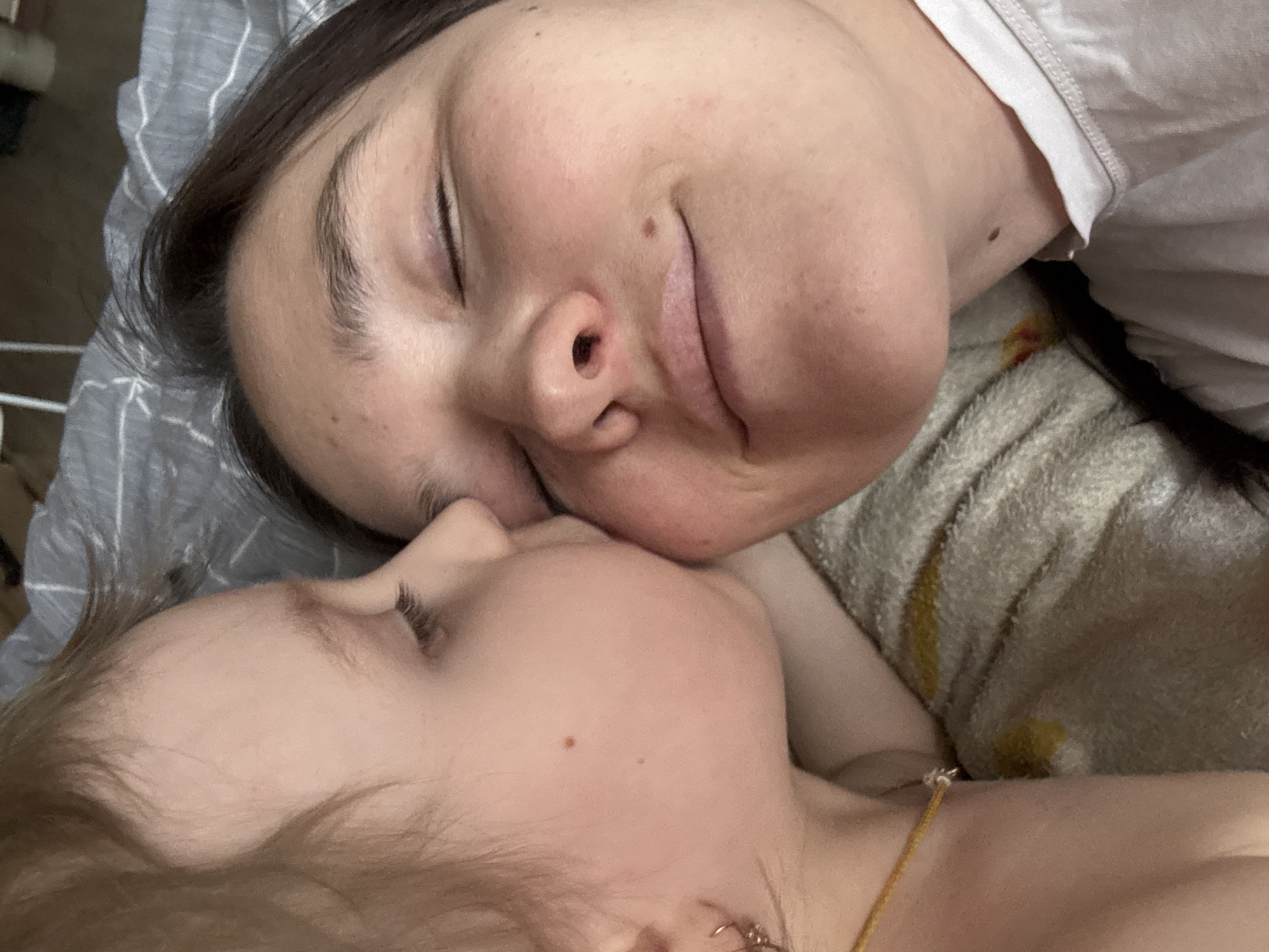This intimate, close-up photograph captures two individuals, likely an adult woman and a young child, lying together in a peaceful indoor setting. The adult, positioned in the upper frame, has dark hair and fair skin, with her eyes closed and a soft, content expression on her face, accentuated by a subtle smile line near her mouth and a mole on her cheek. Her mouth is slightly open, suggesting relaxation or sleep. She has well-defined eyebrows and wears a light-colored, possibly white, top, resting her head on a striped grey and white sheet or blanket.
The child is situated in the lower part of the frame, facing the adult, with their faces almost touching in a tender embrace. The child has lighter hair, possibly blonde, and also has closed eyes with long eyelashes, conveying a sense of calm and sleepiness. A small, light-colored mole is visible on the child's forehead. A thin, light-colored chain or necklace with a small charm is visible around the child's neck.
Both individuals are nestled on soft bedding or blankets; one is striped grey and white, and another light-colored blanket is visible in the background, which shows a small, faint yellowish stain. The lighting is soft and diffused, suggesting an indoor setting during the day, possibly morning or afternoon, enhancing the tranquil atmosphere. The close proximity and relaxed postures evoke feelings of warmth, affection, and security between the two subjects, indicative of a parent-child bond. There is no visible text in the image. The described scene is located in Chaikovsky, Russia.