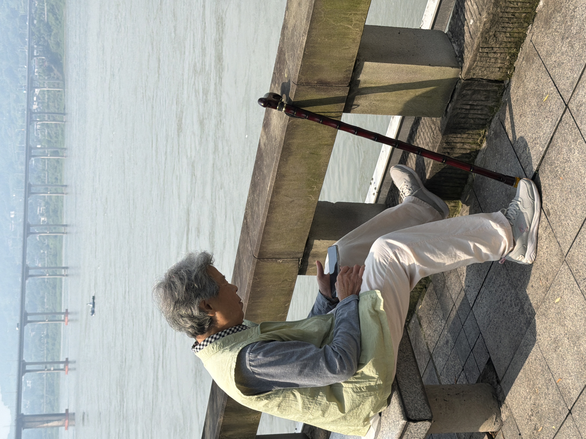 An elderly person with gray hair sits on a stone bench beside a body of water, looking at a smartphone. They are wearing a light green vest over a gray long-sleeved shirt and loose beige pants, with light gray athletic shoes. A dark red cane with a decorative handle rests against a stone railing to the right of the person. In the background, a large bridge spans the water, with a small boat visible in the distance. The scene appears to be a waterfront promenade in Jiangjin, China.