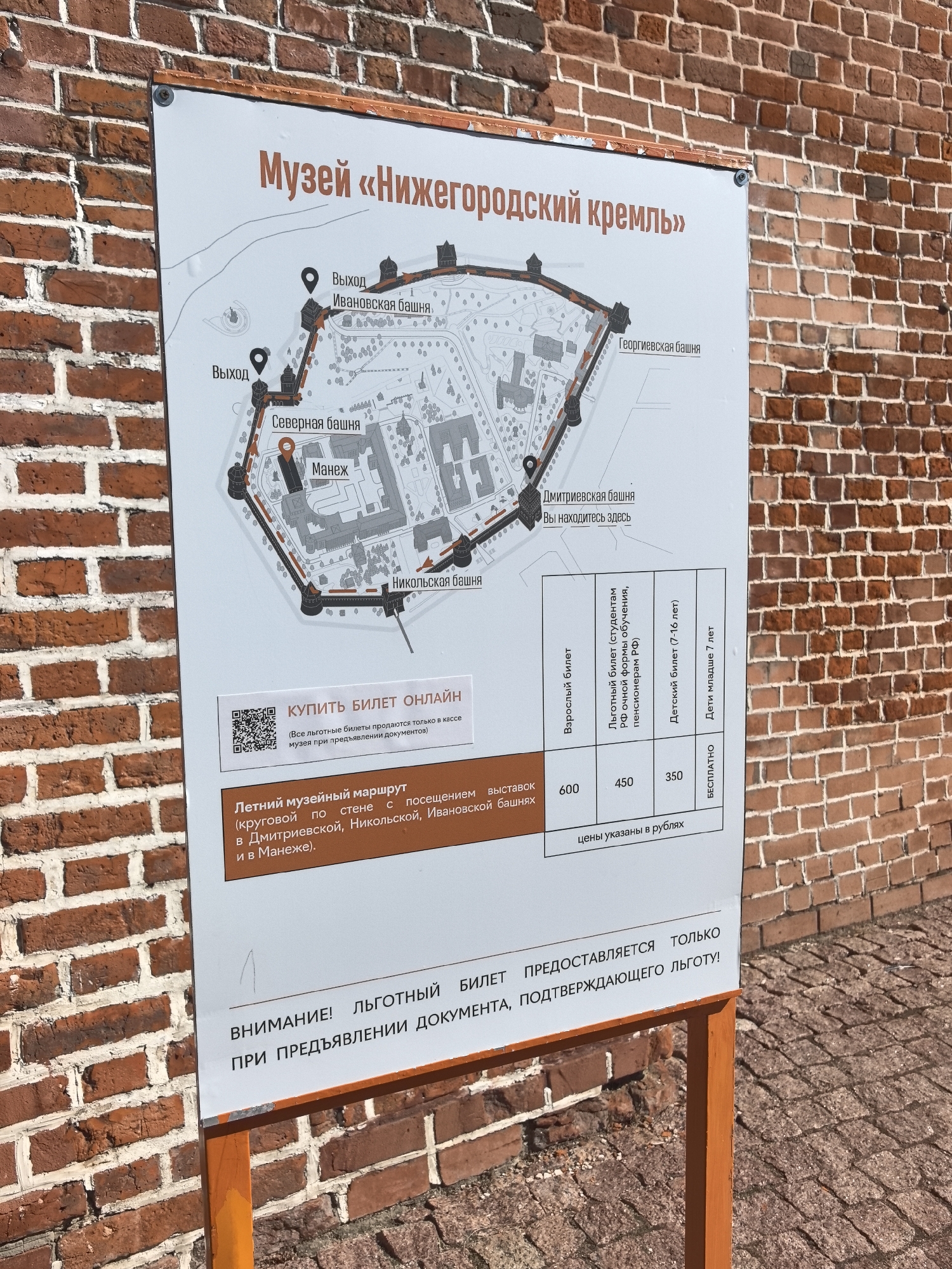 The image shows a sign in front of a red brick wall, likely located in Nizhny Novgorod, Russia. The sign is a map and information board for the "Nizhny Novgorod Kremlin Museum."
The top portion of the sign displays a schematic map of the Kremlin, indicating various towers such as Ivanovskaya, Severnaya, Dmitrievskaya, and Georgievskaya towers. It also shows the location of the "Manège" (an exhibition hall). There are also pointers labeled "Выход" (Exit). One part of the map highlights "Дмитриевская башня" (Dmitrievskaya Tower) with the text "Вы находитесь здесь" (You are here).
Below the map, there is a section with the heading "КУПИТЬ БИЛЕТ ОНЛАЙН" (Buy Ticket Online) and a QR code.
Further down, there is information about a "Летний музейный маршрут" (Summer Museum Route) which includes a circular route along the wall with visits to Dmitrievskaya, Nikolskaya, Ivanovskaya towers, and the Manège.
A table displays ticket prices in rubles:
- Взрослый билет (Adult ticket): 600
- Льготный билет (students, full-time education, RF pensioners) (Concession ticket): 450
- Детский билет (7-16 лет) (Child ticket (7-16 years)): 350
- Дети младше 7 лет (Children under 7 years): БЕСПЛАТНО (Free)
At the bottom, there is a warning: "ВНИМАНИЕ! ЛЬГОТНЫЙ БИЛЕТ ПРЕДОСТАВЛЯЕТСЯ ТОЛЬКО ПРИ ПРЕДЪЯВЛЕНИИ ДОКУМЕНТА, ПОДТВЕРЖДАЮЩЕГО ЛЬГОТУ!" (ATTENTION! A CONCESSION TICKET IS PROVIDED ONLY UPON PRESENTATION OF A DOCUMENT CONFIRMING THE CONCESSION!).
The sign is mounted on an orange metal stand, and the background is a textured red brick wall. The ground appears to be paved with cobblestones. No people are visible in the image.