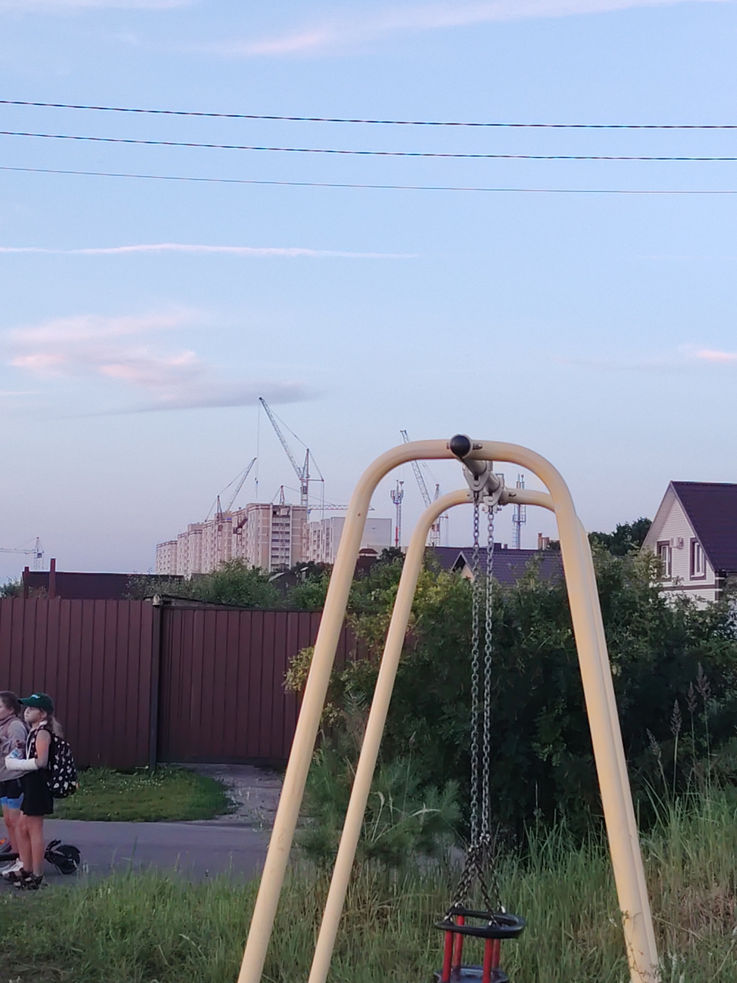 The image depicts an outdoor scene with a playground swing set in the foreground. The swing set has a yellow metal frame and chains suspending a black and red seat. Behind the swing set, a brown wooden fence runs horizontally across the mid-ground. Beyond the fence, there are several tall construction cranes and unfinished buildings, indicating ongoing development in the area. The sky above is a light blue with some wispy clouds and power lines stretched across it. To the left of the fence, a group of three individuals are standing on a paved path, two of whom appear to be young girls. One girl is wearing a black dress with a white polka dot backpack, and the other is wearing a green cap. The environment appears to be a suburban or semi-rural area on the outskirts of the city of Krutye Vyselki, Russia.