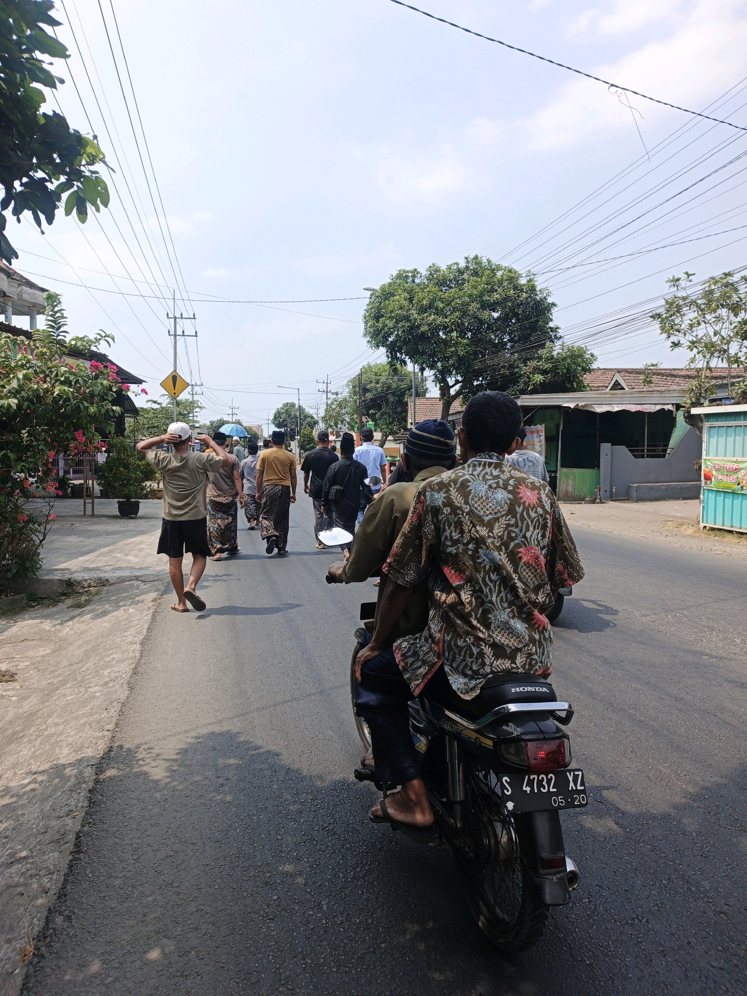 In Mojoduwur, Indonesia, on a sunny day, a group of people are walking on the side of a paved road. In the foreground, two men ride a motorcycle from behind. The man in the back is wearing a batik shirt with a floral pattern and a blue and black cap. The driver is wearing a brown shirt and dark pants. The motorcycle has a license plate that reads "S 4732 XZ 05.20". The street is lined with buildings and trees. In the distance, more people are walking, some wearing traditional clothing. Overhead, power lines are strung across the sky. The overall scene suggests a local event or procession taking place in a village setting.