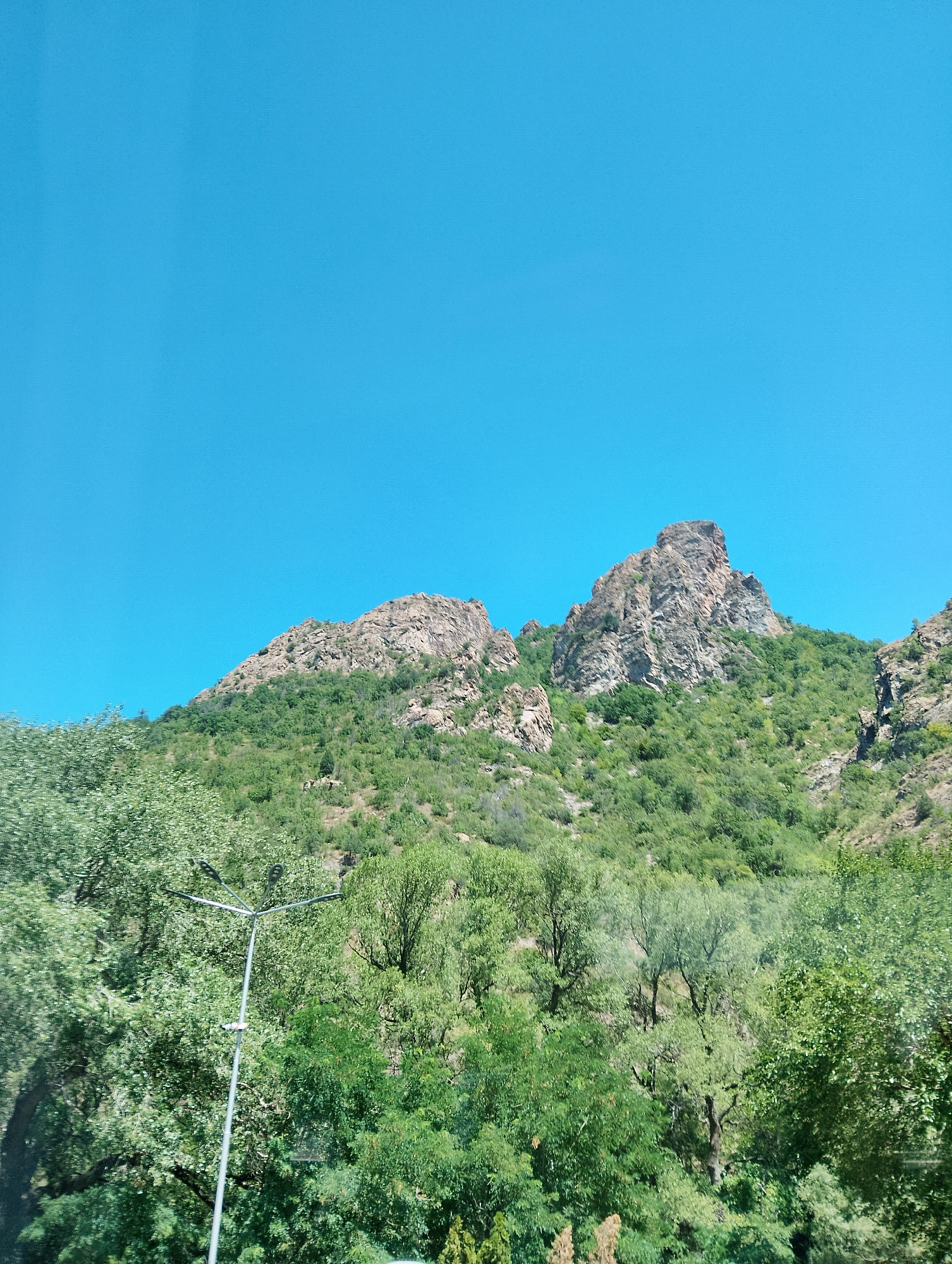 The image captures a bright, clear blue sky above a lush green mountainside covered in trees. Two prominent rocky peaks rise from the center and right of the frame, with rugged, weathered surfaces. The lower slopes are densely forested, with various shades of green foliage indicating different types of trees and shrubs. In the foreground, a silver street lamp with a multi-headed fixture stands tall amongst the trees, its metallic sheen contrasting with the organic textures of the vegetation. The scene appears to be a natural landscape with a man-made element, possibly on the outskirts of a town or city. The image does not show any people or specific actions.