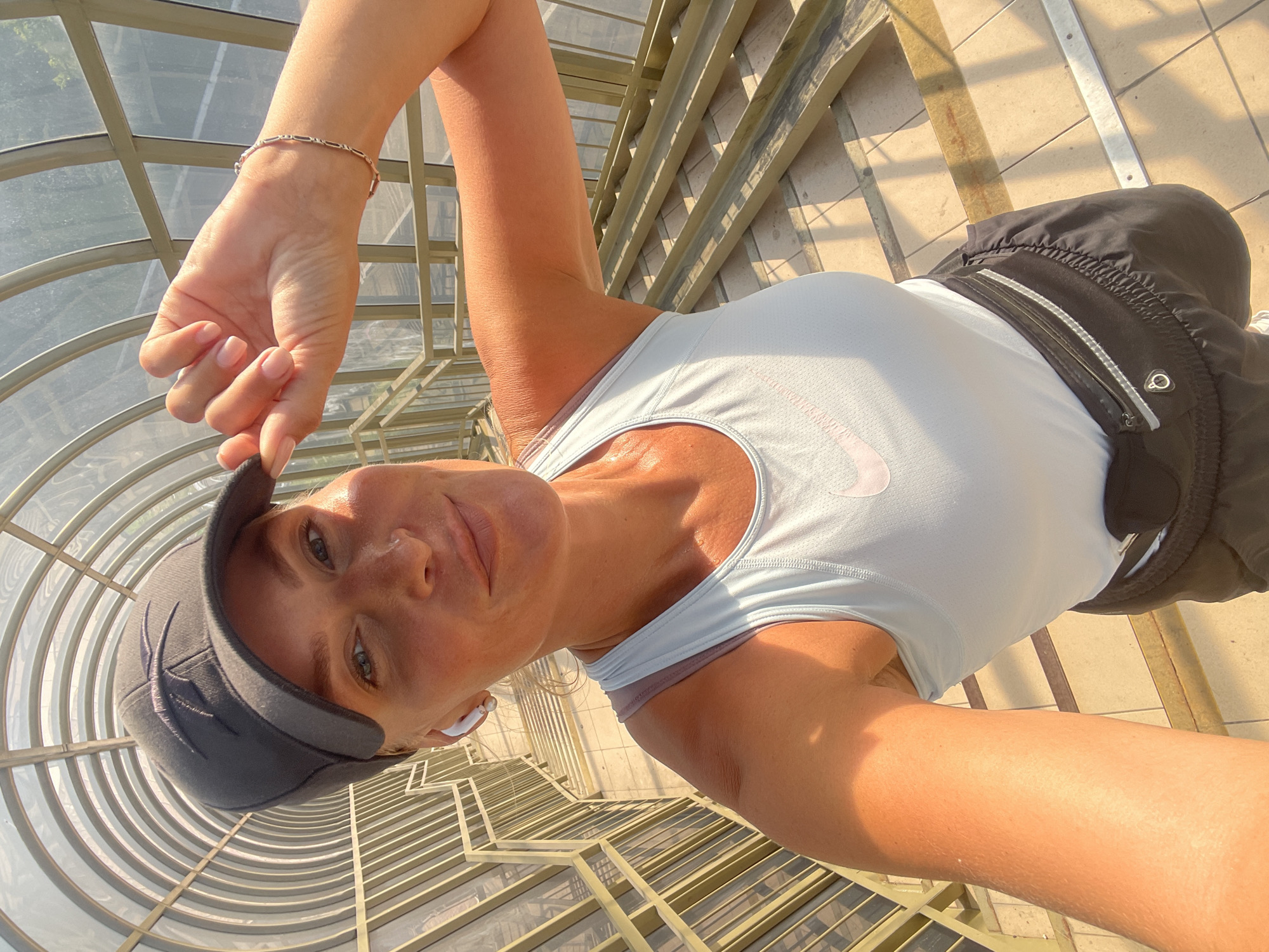 A woman is captured in a selfie taken in an enclosed pedestrian bridge in Sochi, Russia. She is wearing a light blue Nike tank top and black athletic shorts with a fanny pack around her waist. A black baseball cap is on her head, and she is holding the brim with her right hand. She has on white wireless earbuds. The bridge has a curved, transparent roof and metal railings and stairs. Sunlight streams in, illuminating the woman and casting shadows.
