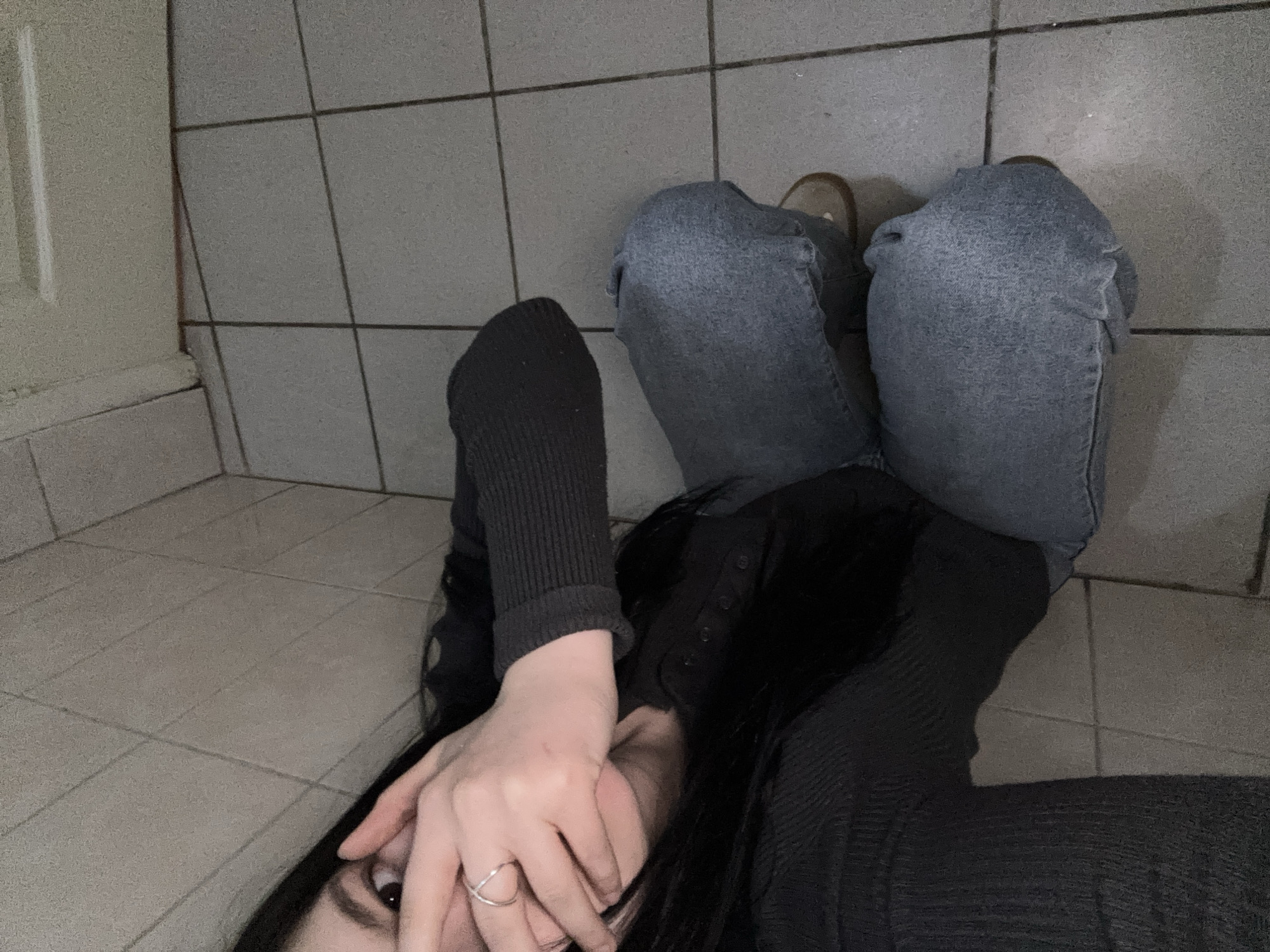 The image depicts a person lying on a tiled floor. They are wearing blue jeans and a dark-colored, possibly black or dark gray, long-sleeved top. The person's hand is near their face, partially obscuring it. Only one eye is visible. A ring is visible on their hand. The tiles suggest the location may be a bathroom or utility area. The background is tiled wall with some other tile on a lower, raised part of the floor. There is a doorframe to the left side of the image.
The location in the city of Chaikovsky, Russia cannot be verified.
