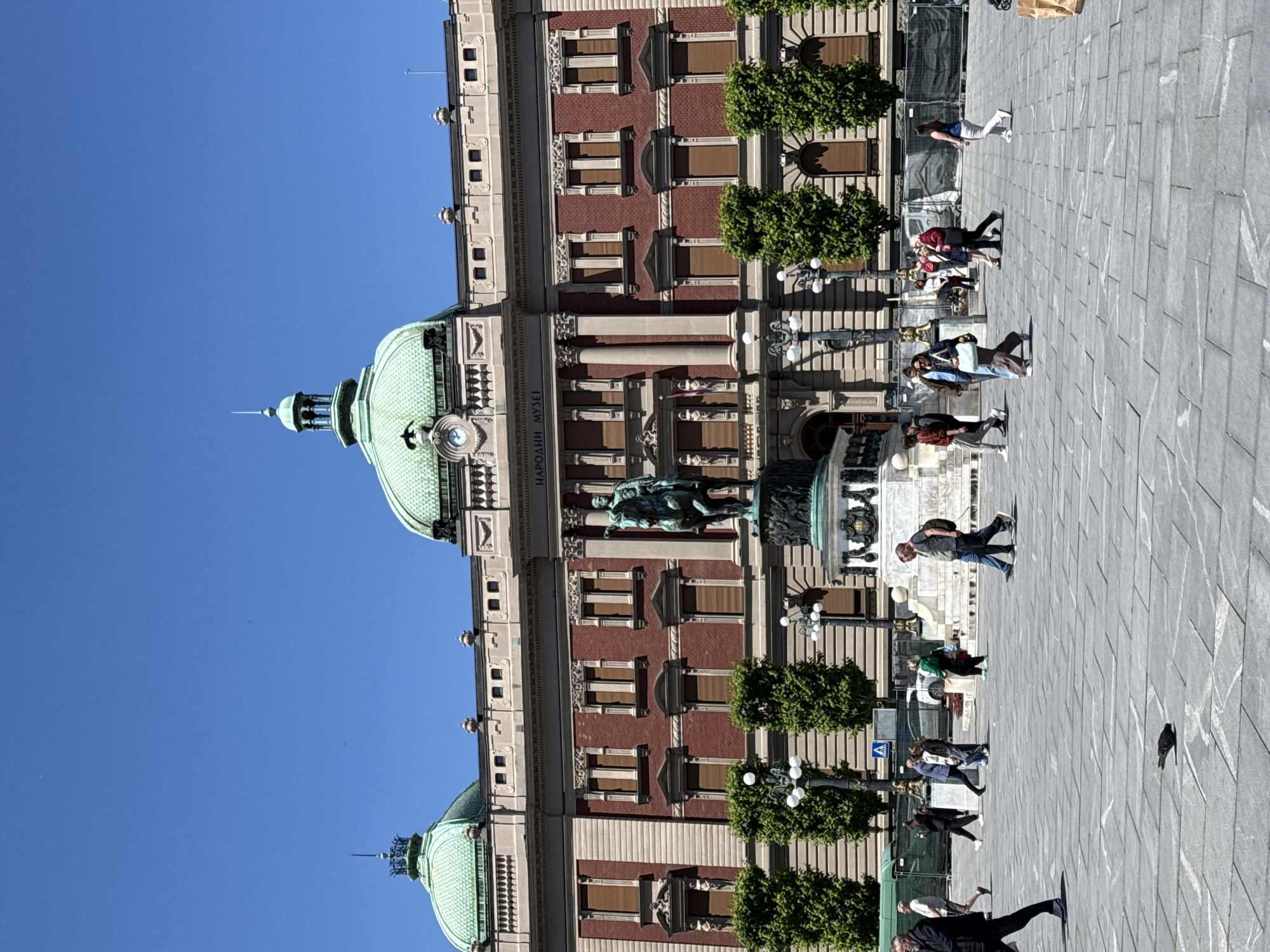 The image depicts the National Museum of Serbia in Belgrade. The museum's facade, constructed of red brick and light-colored stone, features classical architectural elements, including pilasters, arched windows, and a green-domed roof. A statue sits in front of the museum. The surrounding area appears to be a pedestrian square paved with gray stone. Several people are seen walking and standing on the square, along with trees and lamp posts along the facade of the building. The sky is clear and blue.