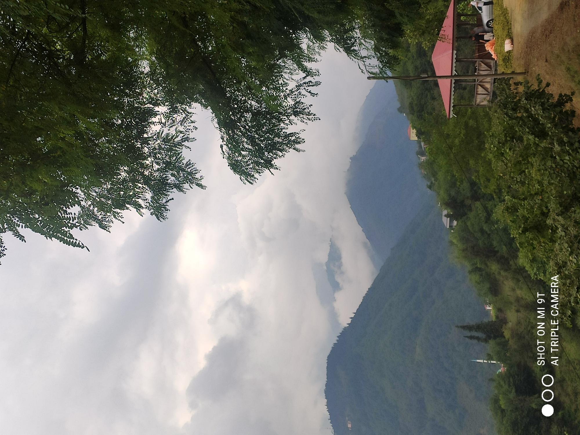 The image captures a panoramic view of a verdant mountainous region, likely a rural area near Akçaabat, Turkey. The foreground features lush green trees, with a prominent cluster of leaves on the right side framing the scene. In the middle ground, a small, rustic gazebo with a red roof is visible. Beneath the gazebo, three individuals are present, one of whom is interacting with a silver car parked next to the structure. Another person is standing near the gazebo. The landscape is characterized by rolling hills and mountains, partially obscured by clouds and mist, creating a serene and atmospheric ambiance. A minaret of a mosque is discernible in the distance on a hillside. The sky is overcast with gray clouds. Text at the bottom of the image reads "SHOT ON MI 9T AI TRIPLE CAMERA."