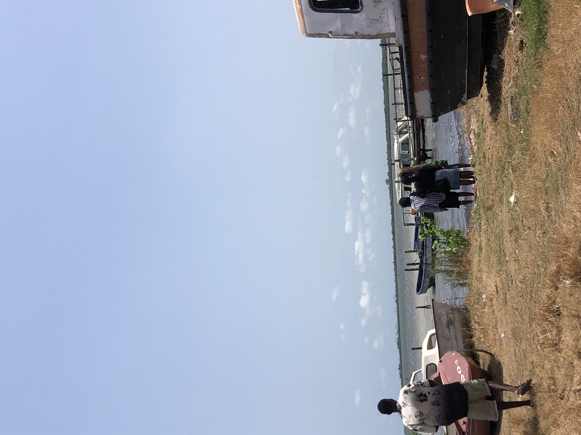 A daytime scene at an unidentified location in Unknown, Unknown, reveals a grassy shoreline adjacent to a body of water. Three individuals stand near a large, partially-viewable truck or vehicle at the water's edge. Additional boats are moored or docked nearby. Another person is walking near an overturned boat on the grassy bank. The sky is pale blue.