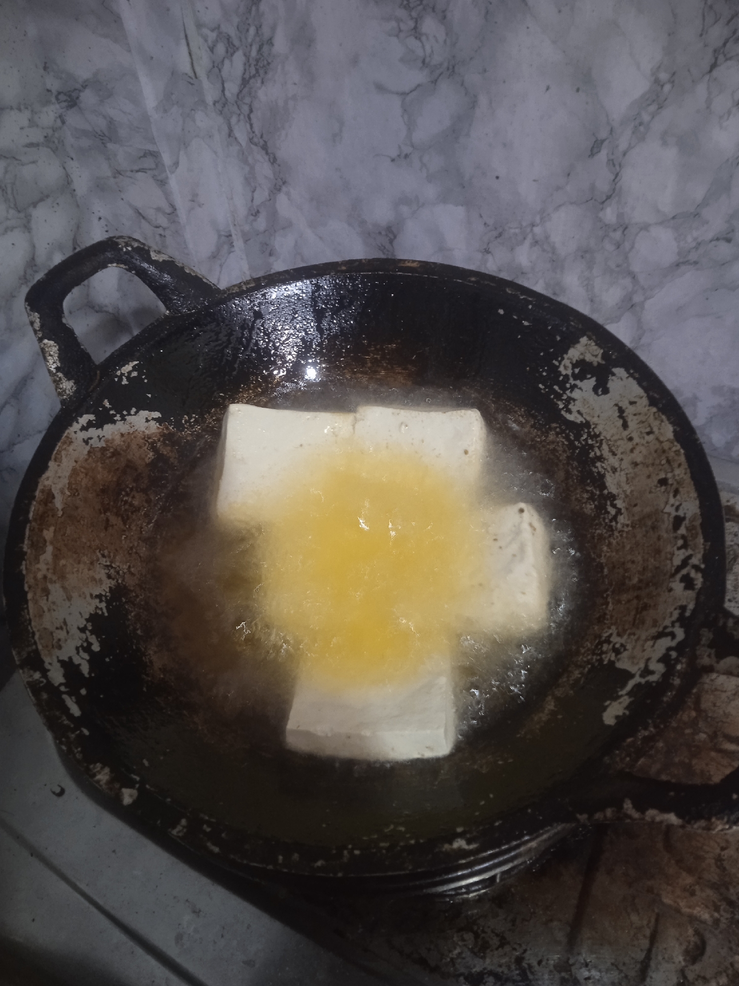 In the city of Surakarta, Indonesia, four white tofu cubes are being fried in a black wok filled with hot oil. The wok is placed on a stovetop, and the oil is bubbling vigorously around the tofu. The background features a marbled wall.