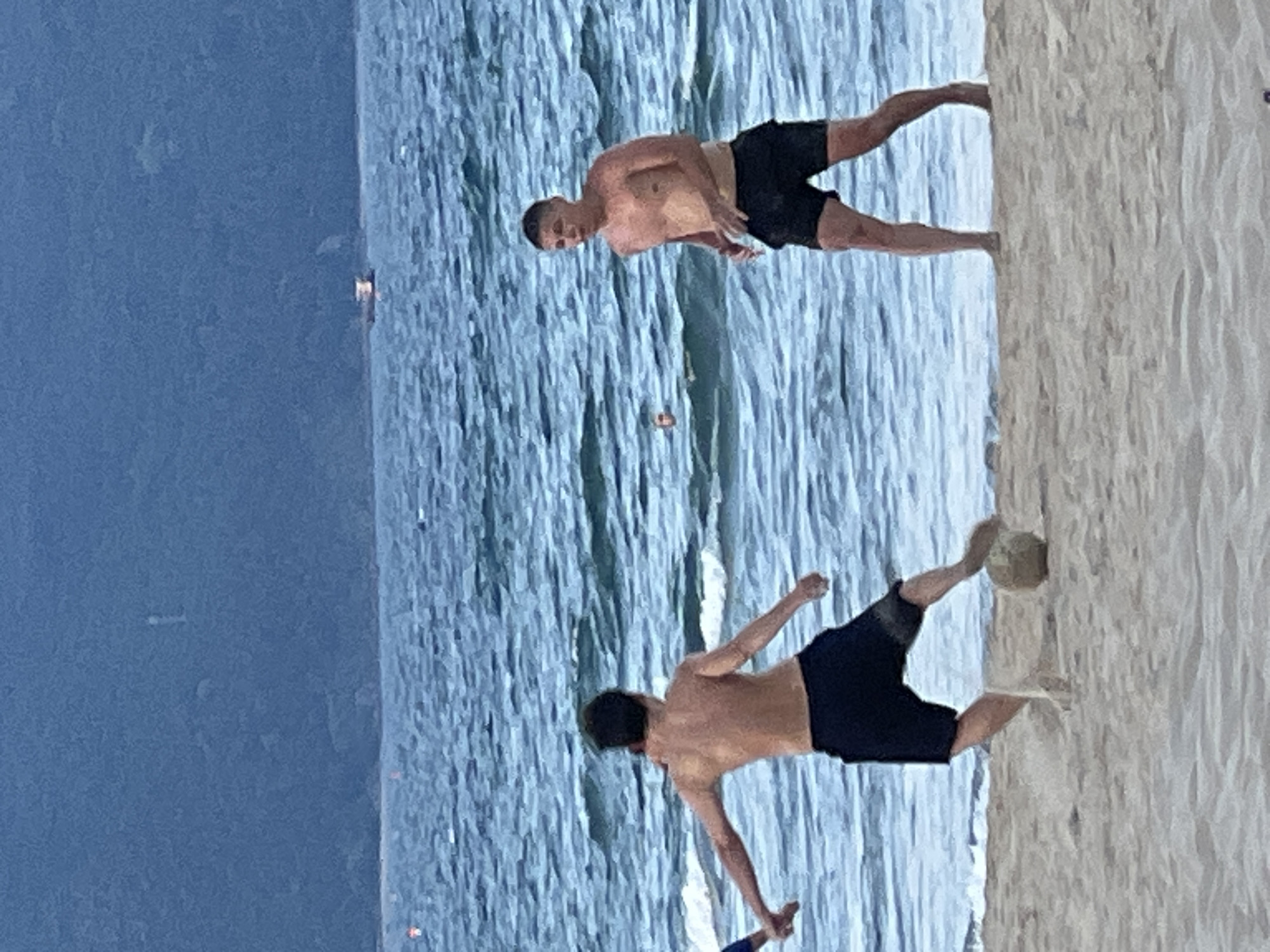 Two men are playing soccer on a sandy beach in Da Nang, Vietnam. The man on the left is kicking a soccer ball, while the man on the right is watching him. The ocean is in the background, with waves and a distant boat. The sky is overcast.