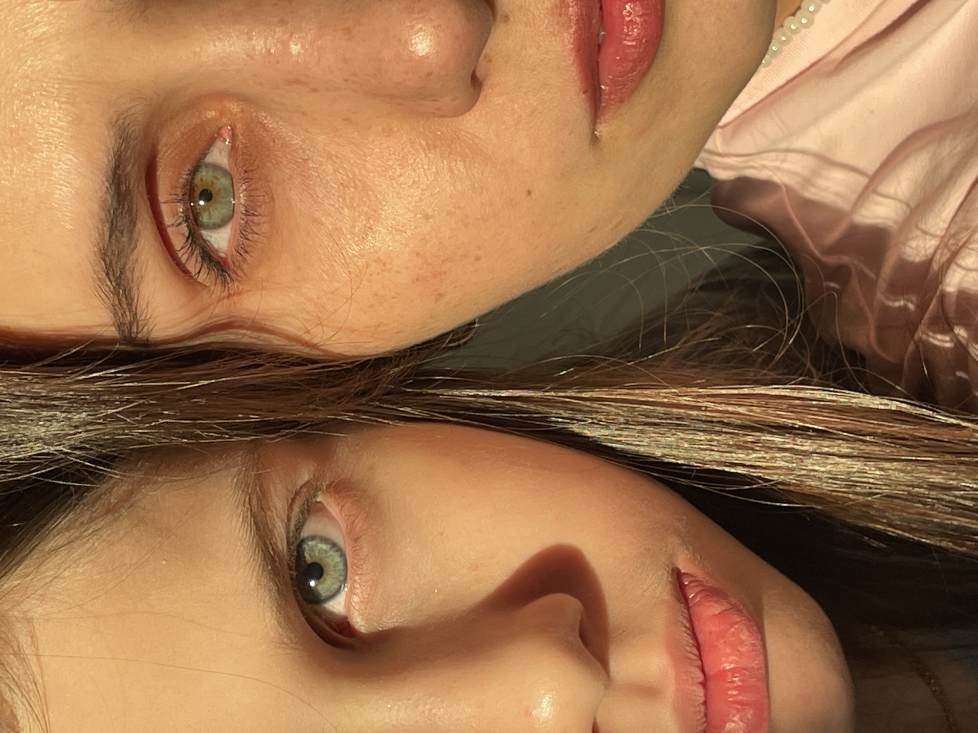 The image displays a close-up, rotated view of two individuals' faces, likely two women or a woman and a child, positioned side-by-side and facing each other. They appear to be lying down, with their heads very close, suggesting intimacy or a close relationship.
The top individual (when viewing the image in its original orientation) has a visible left eye of a striking green-hazel color, framed by dark, well-defined eyebrows and long eyelashes. Her skin is fair, with noticeable freckles scattered across her cheek and around her nose. Part of her nose and her glossy, reddish-pink lips are visible. Her hair, dark brown, flows downwards. Near her ear, a soft pink garment, possibly a shirt or pajama top, is visible, adorned with a delicate trim of small, white, pearl-like beads.
The bottom individual, mirrored in pose to the top, also shows her left green-hazel eye, which appears slightly lighter than the first. Her eyebrows are a softer, lighter brown. Her skin is fair and smooth. Her lips are full and glossy with a similar reddish-pink hue. Her dark brown hair features prominent lighter brown or golden highlights and several thin, shimmering, glitter-like strands interwoven throughout, giving it a luminous quality. This hair is spread out, forming a backdrop and flowing between the two subjects.
The lighting is soft and warm, creating gentle highlights on their skin and glossy lips, and casting subtle shadows that define their facial contours. The overall impression is one of a peaceful and affectionate close-up portrait, capturing their similar features and connection. There is no visible text in the image.