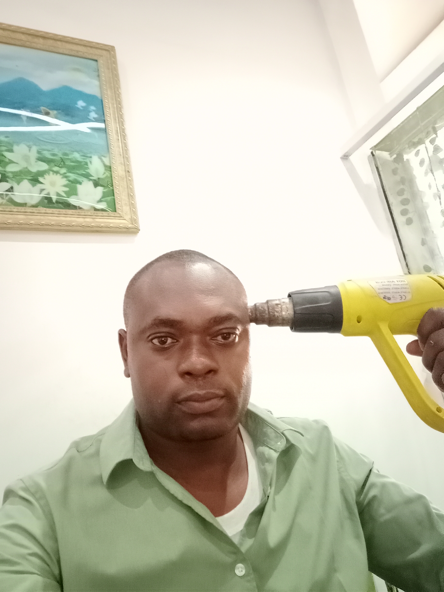 A man is holding a yellow heat gun to his forehead. The man is dark-skinned, bald, and wearing a light green button-down shirt. He is in a room with a picture frame hanging on the wall behind him and a window to the right. The location is Gwarinpa, Nigeria.