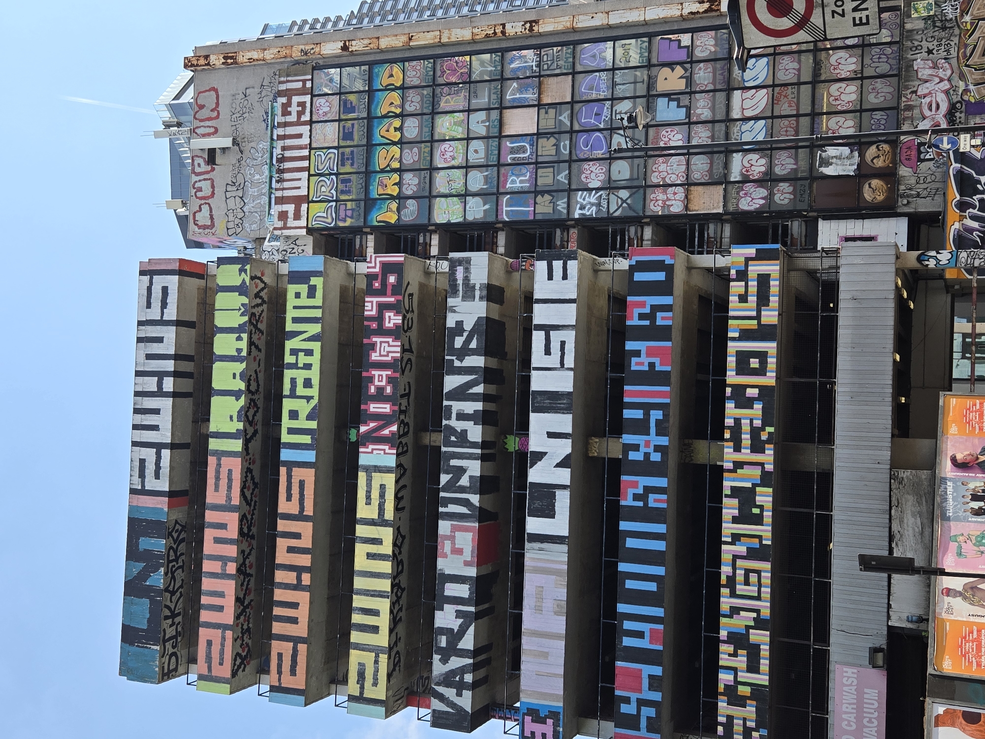 This is a low-angle shot of the Seven Sisters Car Park, a building renowned for its street art in the Finsbury Park area of London, United Kingdom. The building has a partially open facade made of concrete and steel and is covered in graffiti and painted artwork. The top portion has a grid-like window design with painted letters and tags. Underneath this section, vertical concrete beams are brightly painted with stylized text. The bottom portion of the building is covered with advertisements and additional graffiti. The sky is visible above the building.