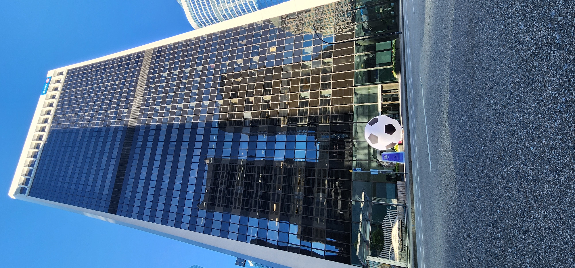 A low-angle, full shot captures a large, modern skyscraper against a clear blue sky. The building's facade is predominantly dark glass, reflecting the surrounding environment and other buildings. A white trim accents the top of the skyscraper, with a visible logo that appears to read "BMO". In the foreground, a massive, inflatable soccer ball is positioned on the sidewalk in front of the building. To the left of the soccer ball, there is a bus stop shelter with a bench. The street in front of the building is paved, with visible white lane markings. The overall scene suggests an urban setting on a bright, sunny day.