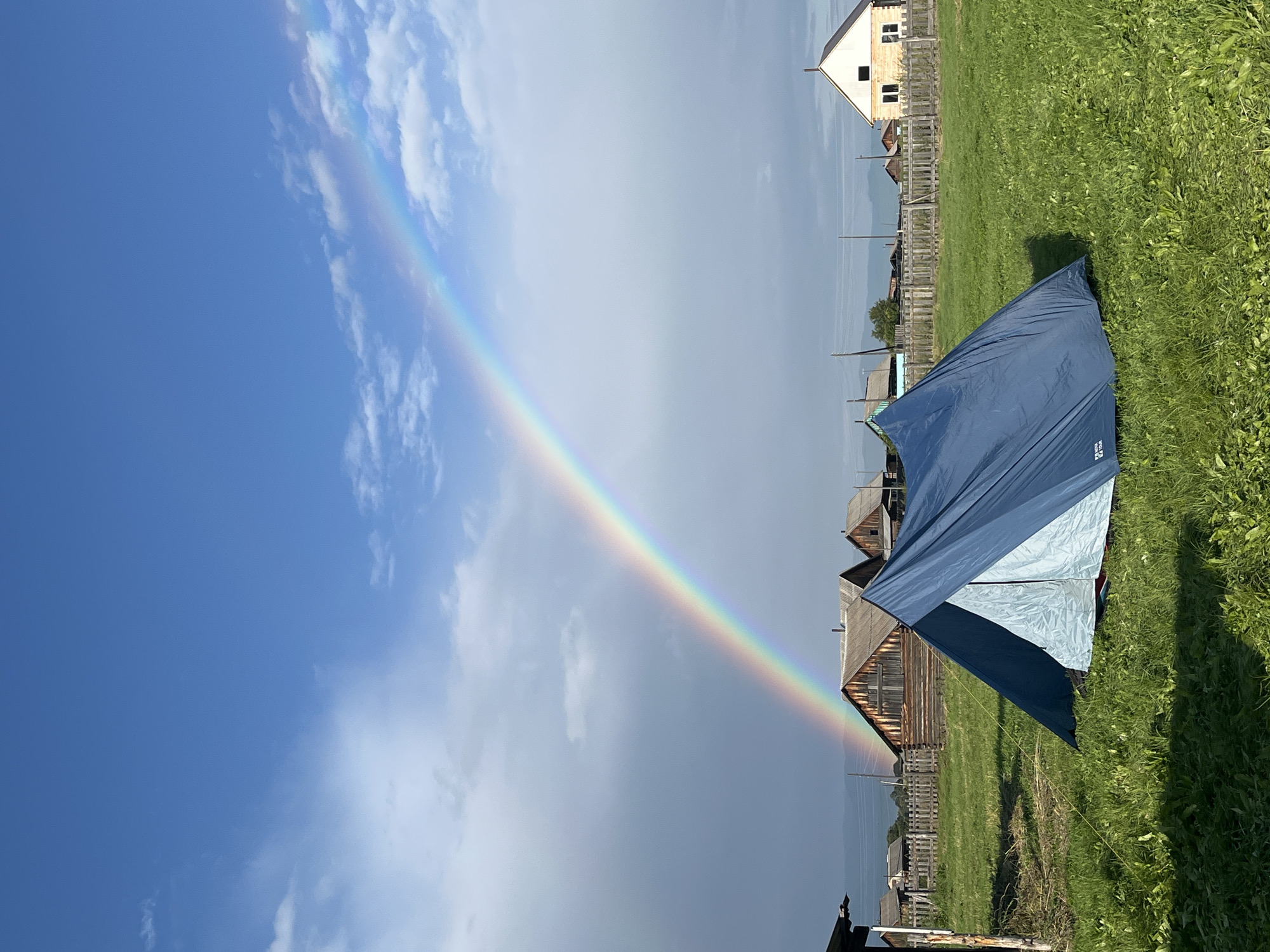 In a grassy field, a blue tent is pitched. Behind the tent, a wooden fence separates the field from a village with several wooden houses and one white house with yellow trim. In the sky above, a bright rainbow arcs across a blue sky with scattered white clouds. The location is Unknown, Unknown.