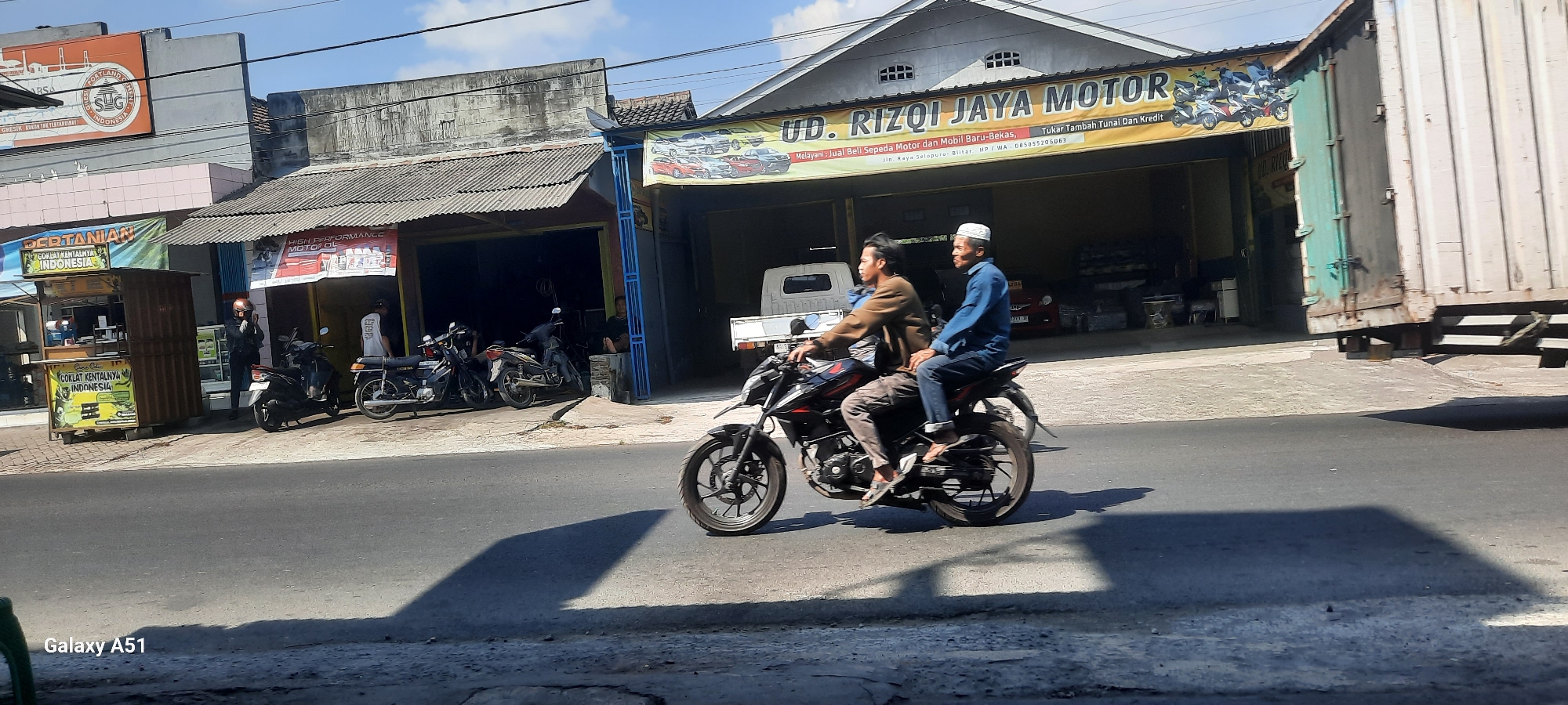 The image captures a street scene in Selopuro, Indonesia, on a sunny day with a clear blue sky. The foreground features a paved road with a distinct shadow of a motorcycle and its riders cast across it. Two individuals are prominently riding a black motorcycle in the middle ground; the rider in front wears a brown jacket and patterned pants, while the passenger behind wears a blue long-sleeved shirt and jeans, with a white skullcap on his head.
Behind the motorcycle, on the left side of the image, is a row of buildings. The closest building has a corrugated metal roof and houses a stall displaying banners advertising "COKLAT KENTALNYA INDONESIA" (Indonesian Condensed Chocolate). A person in a dark jacket and helmet stands near this stall. Further down the row, another building has a similar roof and a banner overhead that reads "UD. RIZQO JAYA MOTOR" (Rizqo Jaya Motor Company), advertising motor and car sales, including options for cash or credit purchases. Several motorcycles are parked outside this establishment. A white truck is visible parked in the garage of the motor company.
To the right of the motor company, a large, weathered, greenish-blue shipping container is partially visible. The overall environment appears to be a commercial area along a road, likely in a small town or city.