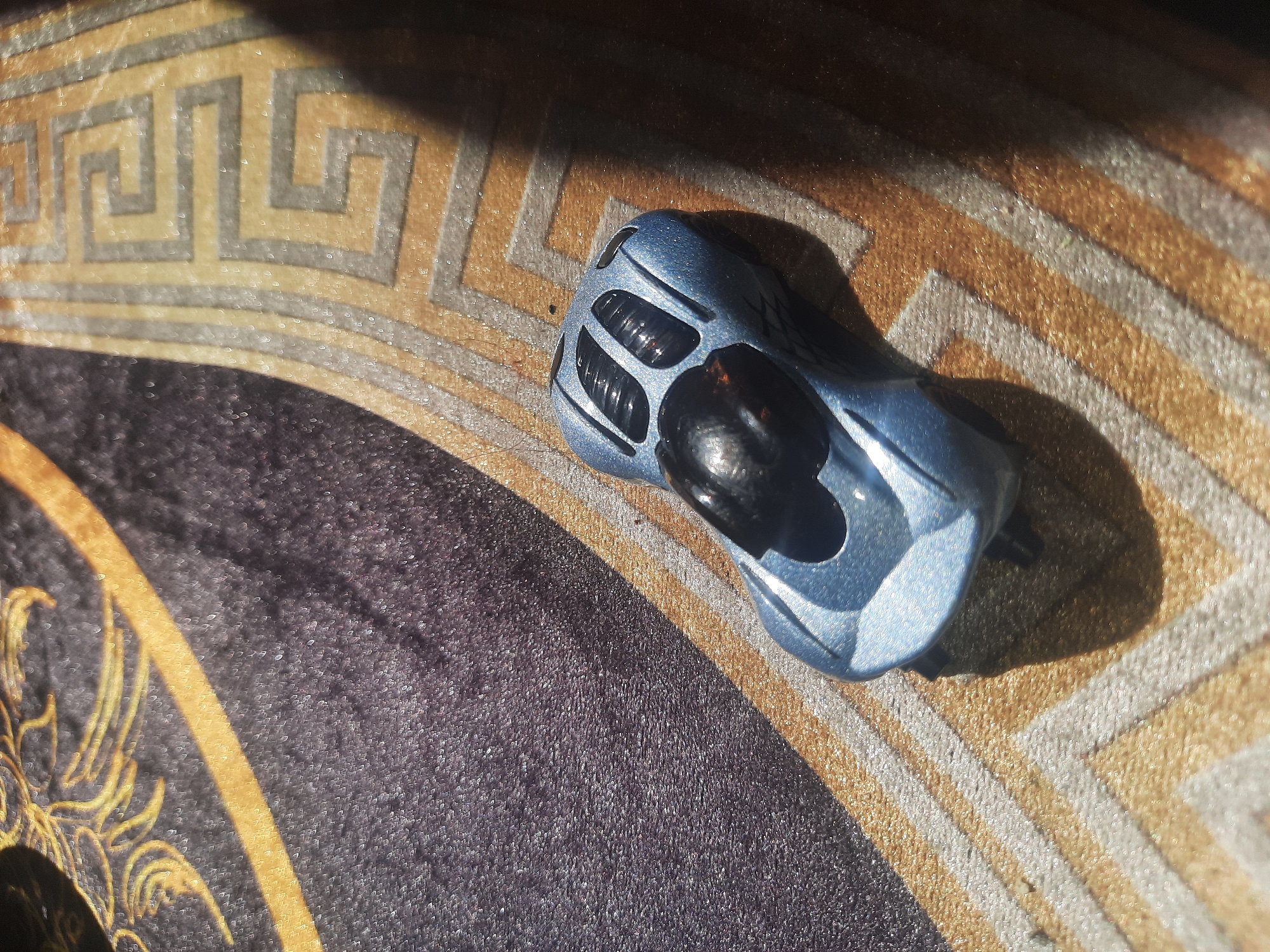 A light blue toy race car is positioned on a patterned rug. The rug features a geometric design with bands of gold and grey against a darker background. A shadow falls across the right side of the image, obscuring part of the rug and car. The scene is set indoors, likely in a residential environment. The specific location in Shklov, Belarus cannot be determined from the visual information provided.