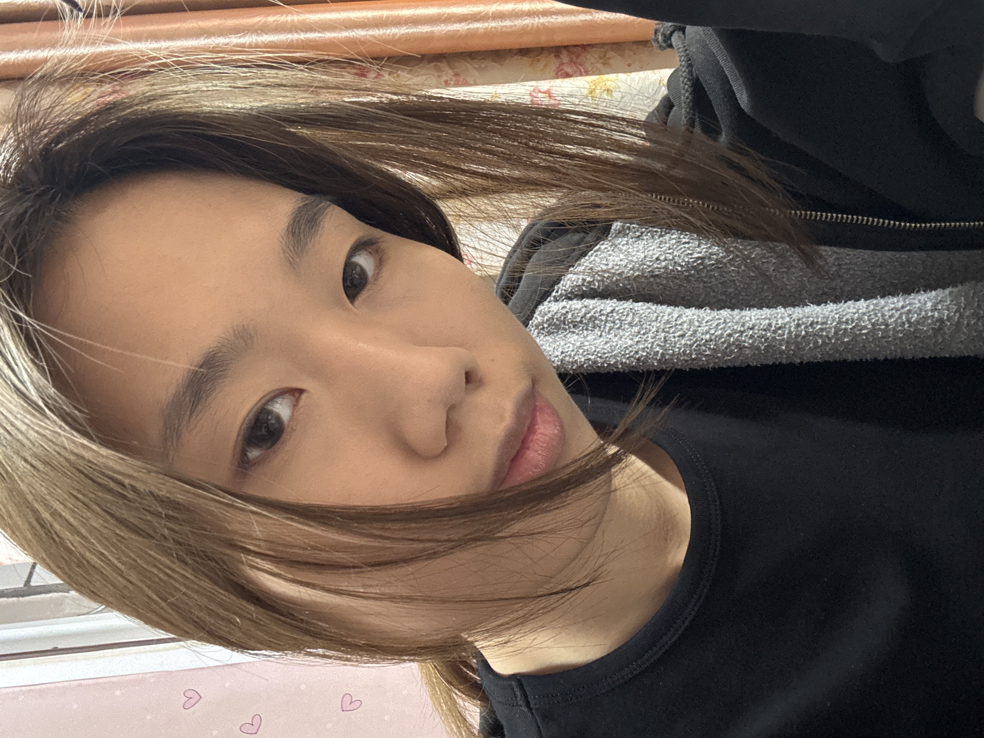 A close-up, angled selfie captures a young woman with light brown hair partially obscuring her face. She has brown eyes and is wearing a black, crew-neck top layered with a gray and black zippered hoodie. The background is blurry, but reveals a floral patterned surface and what appears to be a window with white blinds. The presence of a blanket with a heart pattern further back suggests an indoor setting, possibly a bedroom. It is not possible to confirm the location of the Shapingba District, China based on the visual elements of the image alone.