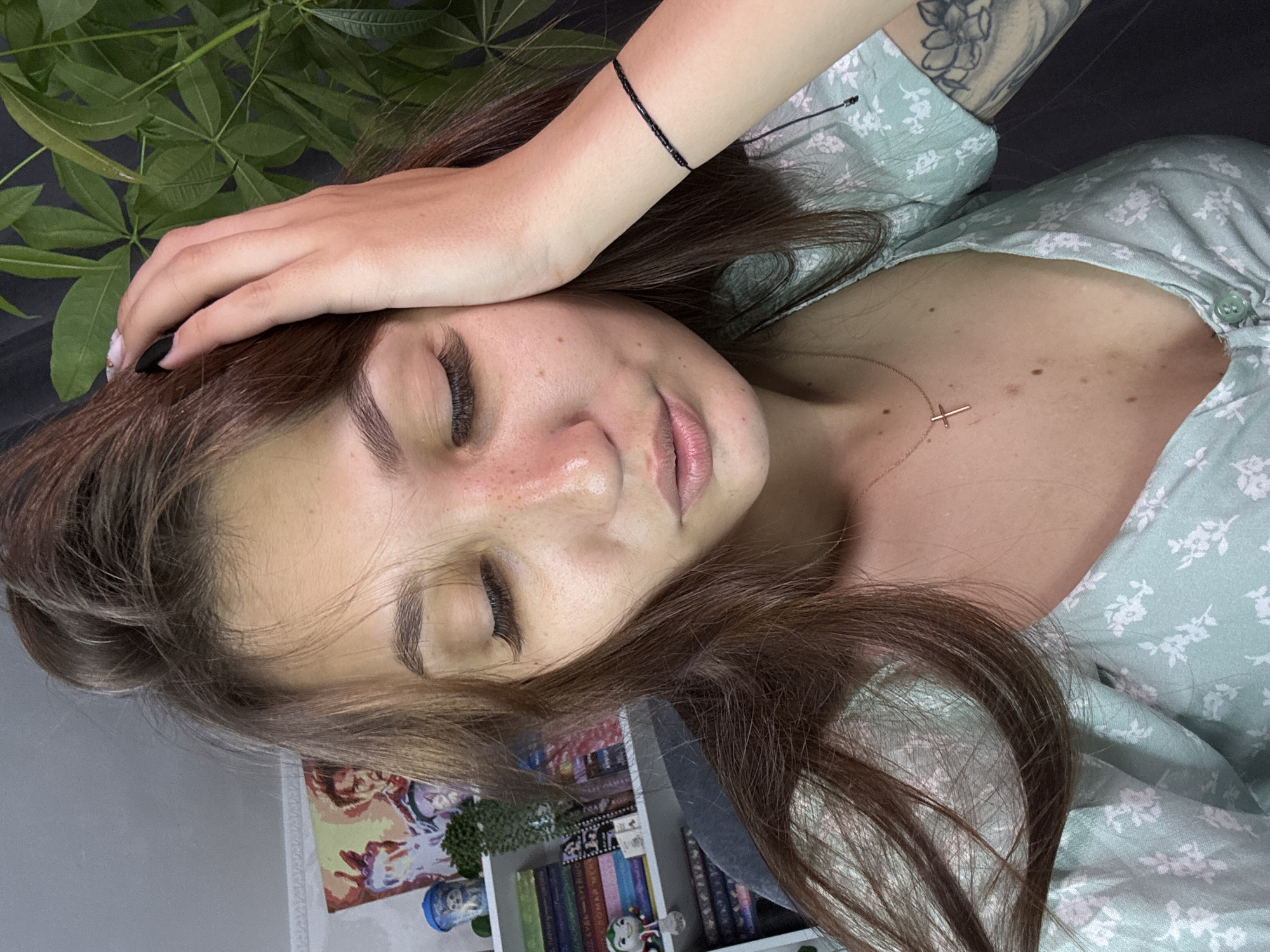 A young woman with long brown hair, wearing a light green floral top, is captured in a close-up, overhead shot. She is resting her head on her hand, eyes closed, with her lips slightly parted. Her face shows some freckles and light blushing across her cheeks. She is wearing a delicate gold cross necklace and a black beaded bracelet. In the background, a bookshelf filled with books is visible, along with a potted plant. The image is taken indoors, likely in a home setting. No specific location in Novokuznetsk, Russia can be determined from the image.
