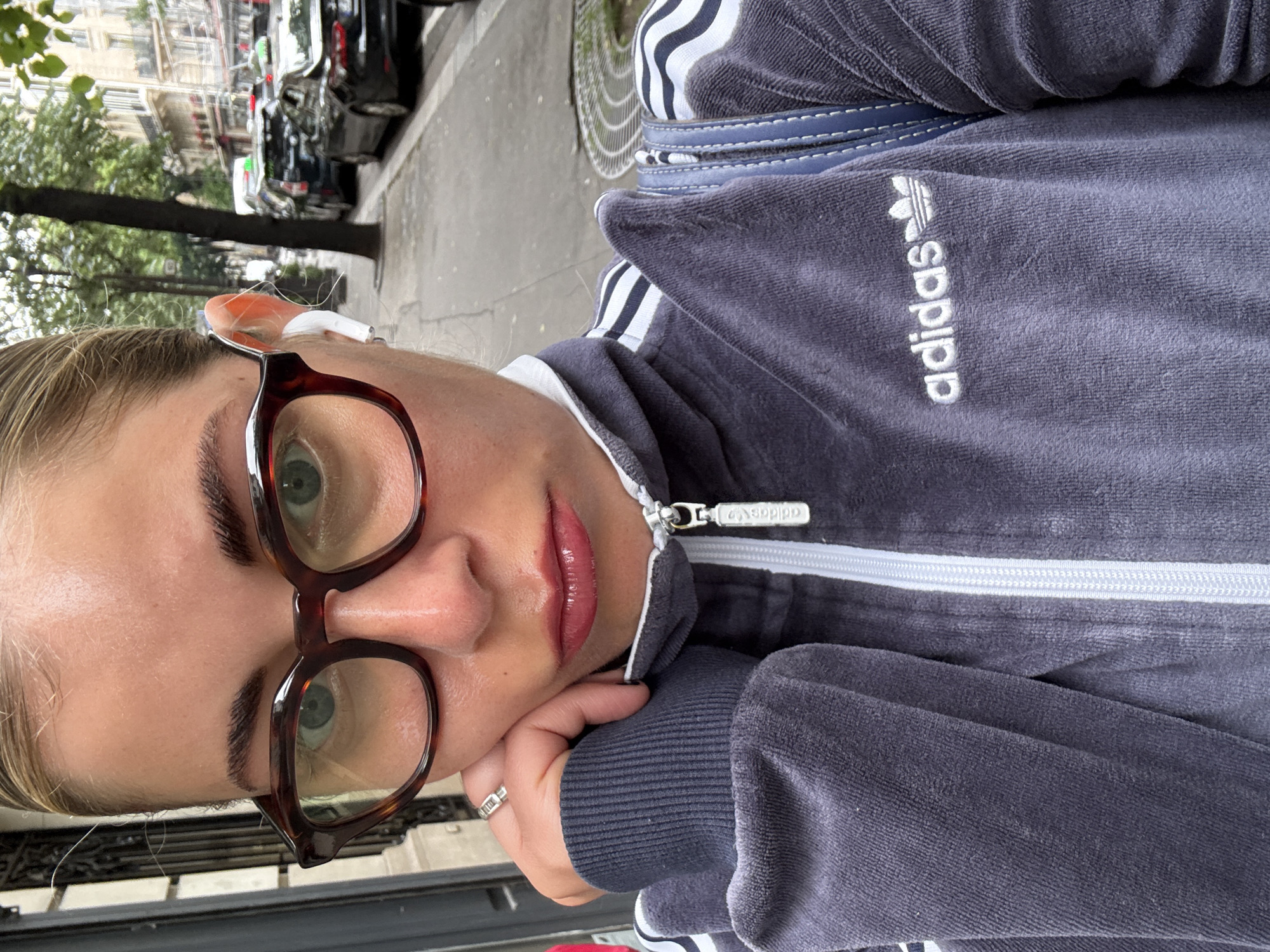 A woman with blonde hair and green eyes is photographed in what appears to be Paris, France. She is wearing brown-rimmed glasses and an Adidas tracksuit with the brand's logo visible. She has an earpiece in her ear. The background shows a street with parked cars and trees, characteristic of a Parisian cityscape.
