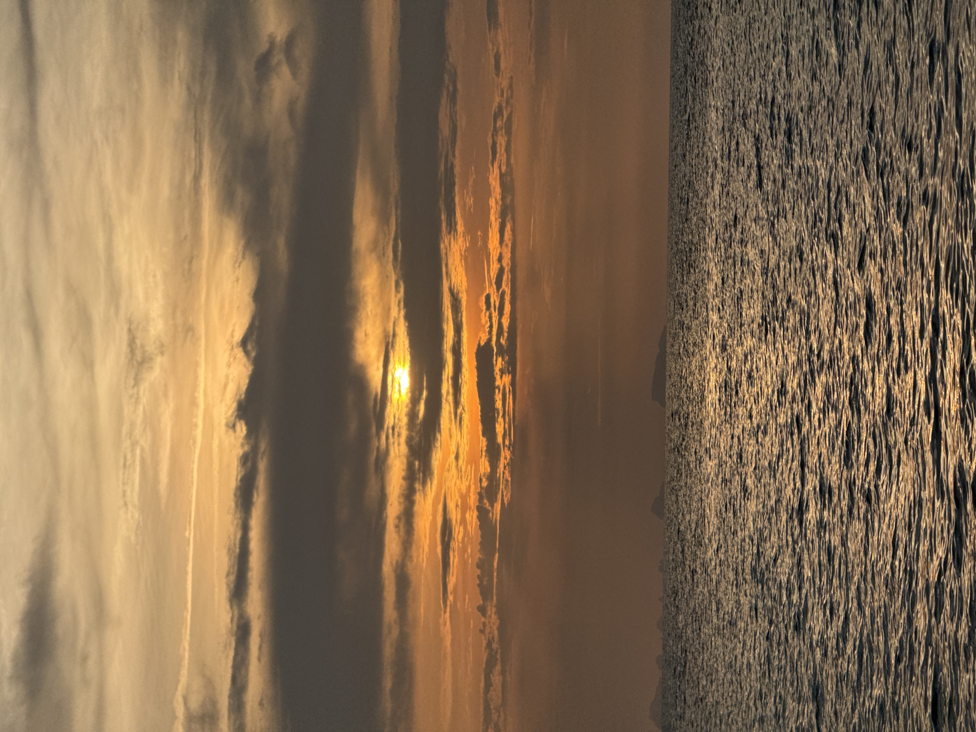 A vertical, medium shot captures the expanse of the ocean at sunset in Ko Samui District, Thailand. The sun, partially obscured by thick, horizontal bands of dark clouds, casts an orange glow across the sky. The water, a deep, shimmering blue, is textured with small waves that reflect the warm light. In the distance, silhouetted islands appear as faint, dark shapes against the hazy horizon. The scene is devoid of any people or artificial structures, emphasizing the natural beauty of the seascape.
