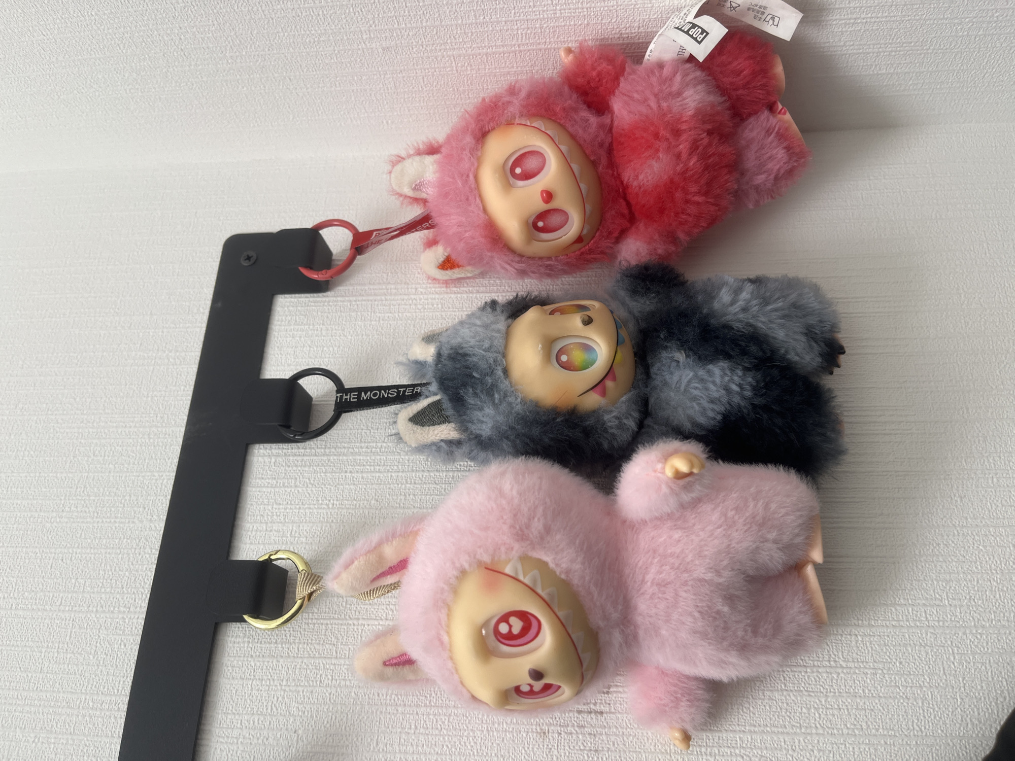 Three plush keychains are displayed hanging from a black metal wall-mounted rack against a white textured wallpaper in Saint Petersburg, Russia. From left to right: The first keychain is a pink, fluffy figure with a bunny-eared hood, featuring a pale, stylized face with a wide, toothy smile and large pink eyes with heart-shaped pupils. It is suspended by a gold-colored keyring. The central keychain is a grey and black fluffy figure with animal-like ears, a pale face displaying a toothy grin, and large eyes with multi-colored, rainbow irises. This figure hangs from a black keyring, which has "THE MONSTERS" printed on its strap. The third keychain, on the right, is a reddish-pink fluffy figure, also with a bunny-eared hood, a pale face, a wide, toothy smile, and large pink eyes. It is attached to a red-colored keyring. All three figures are presented vertically, occupying the upper-middle portion of the frame.