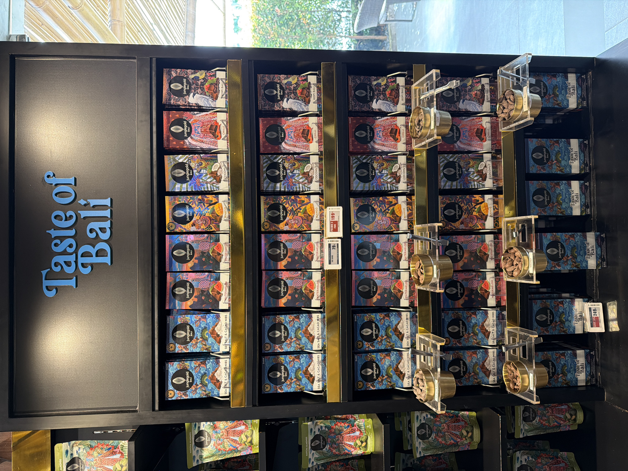 A photograph taken indoors shows a display shelf filled with neatly arranged products, predominantly chocolate bars. The words "Taste of Bali" are displayed in blue on the left side of the shelf. The chocolate bars are branded "Junglegold Bali" and feature various flavors, including Kopi Bali, Chilli, Coconut, Mango, Watermelon, and Sea Salt & Cacao Nibs. The display includes promotional tags, like "Promo 2 For 199K." Clear acrylic holders with small bowls filled with what appears to be samples or ingredients are placed alongside the chocolate bars. The shelf is black with gold dividers. The top of the shelf is near a window with views of exterior bamboo shoots and greenery, suggesting an indoor environment, possibly a shop located in Mekarsari, Indonesia.