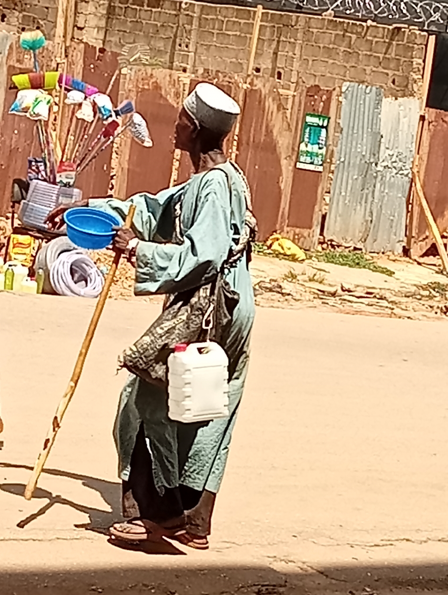 In Maiburiji, Nigeria, an elderly man is walking along a paved road. He is wearing a light blue robe, a white cap, and sandals. He holds a wooden staff in his left hand and a blue bowl in his right. A white plastic container hangs from his shoulder. Behind him is a cart laden with household items, including brooms and plastic containers. The background shows a brick wall with corrugated metal sheets.