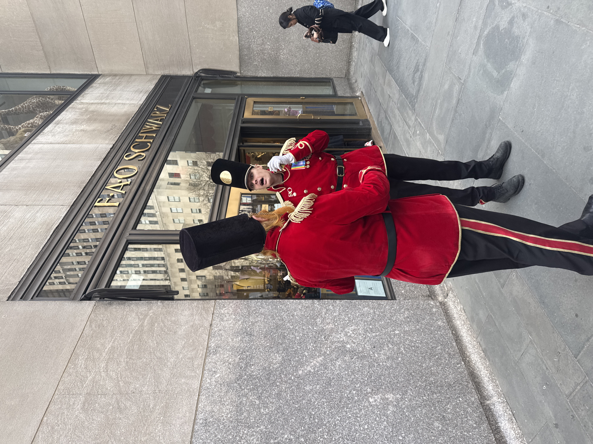 The image captures a street scene in front of the FAO Schwarz toy store at Rockefeller Center, New York City, United States of America. Two individuals, dressed in elaborate red and black toy soldier uniforms, stand on a light grey flagstone sidewalk. Each uniform features a red tunic with gold trim and epaulets, black trousers with a prominent red and gold stripe down the side, and a tall black shako (hat) adorned with a gold medallion.
The individual positioned to the left is viewed from behind, with reddish-blonde hair visible beneath their shako. The individual to the right faces the first, their mouth open in an expression suggestive of speaking or singing, while a white-gloved hand is held near their chin. This individual's uniform displays multiple colorful ribbons and a medallion on the left breast.
Behind these figures, the storefront of FAO Schwarz is visible, featuring large glass doors serving as the entrance and extensive display windows. Gold-colored block lettering spelling "FAO SCHWARZ" is affixed above the entrance. Through the upper windows, the heads and necks of large stuffed giraffe figures are discernible. The glass surfaces also reflect surrounding city buildings.
To the far right of the frame, a woman dressed in dark clothing (black top, black pants, white sneakers) walks away from the viewer on the sidewalk, holding a brown handbag and a blue tote bag. She appears to be looking down at an object in her hand. The building's facade consists of textured, light-colored stone panels above a darker, polished stone base.
