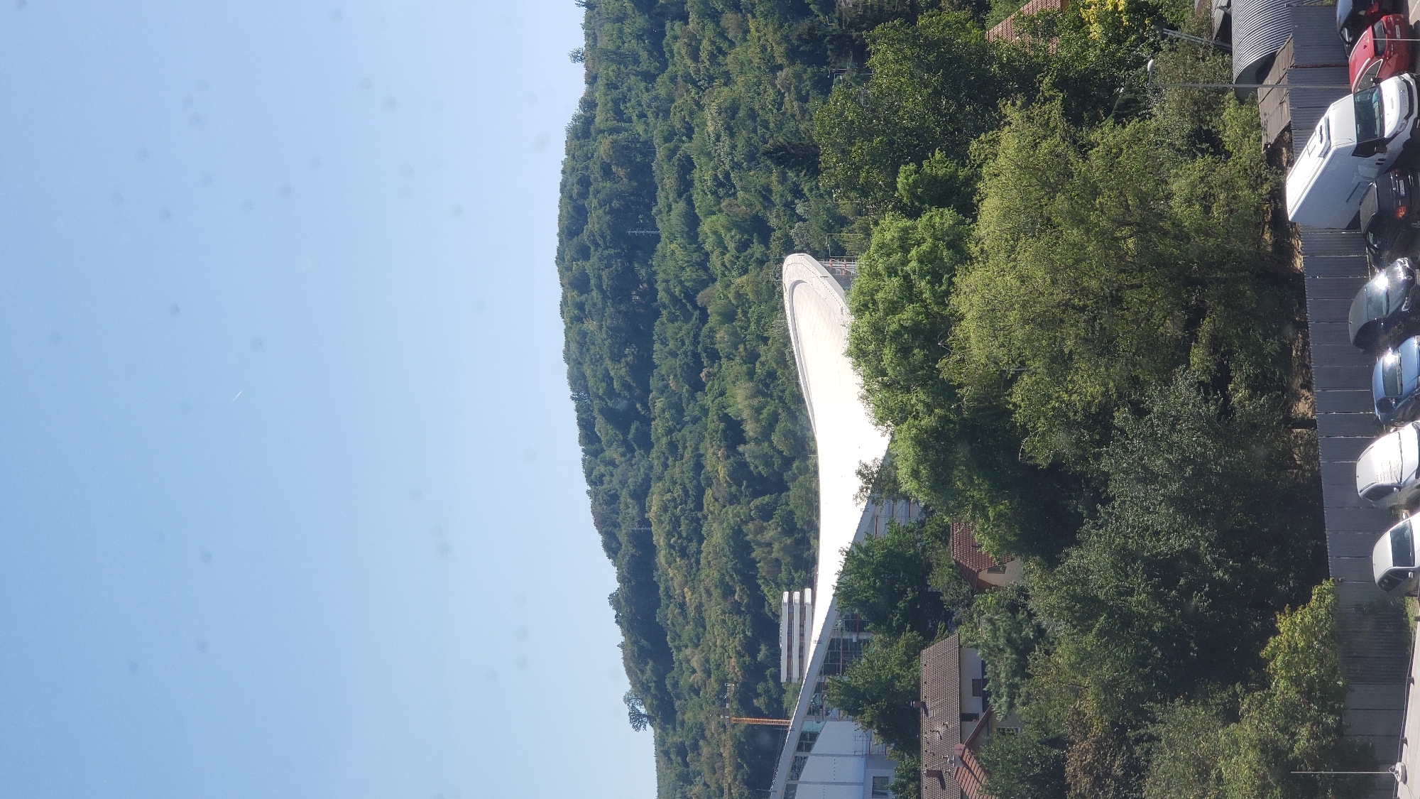 The image shows a view of the ski jump at the Summer Ski Jumping Center in Prešov, Slovakia. The ski jump's white ramp is prominently featured against a backdrop of a densely wooded hill. Several trees are also visible in the foreground, along with some residential buildings. In the lower part of the image, a row of parked vehicles, including cars and a white van, are visible behind a metal fence. The sky is clear and blue, with a faint contrail from an airplane visible. The scene appears to be set on a bright, sunny day.