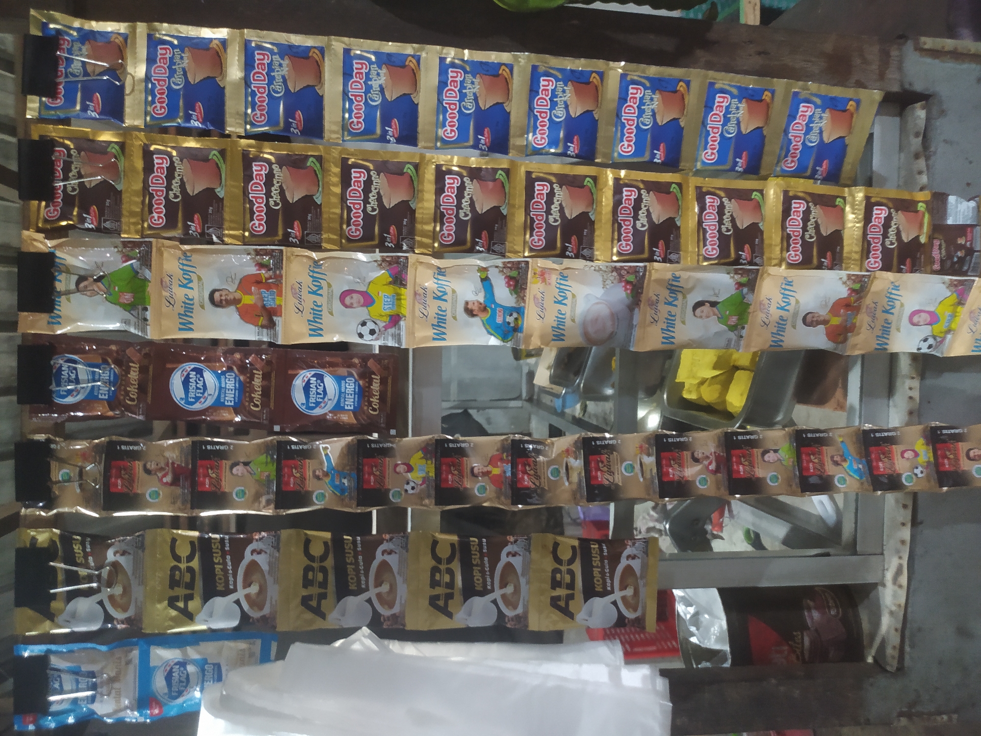 The image shows a street-side stall in Tuntang, Indonesia, displaying an assortment of packaged coffee and milk products. The stall features multiple rows of colorful sachets hanging from a rack. The majority of the products are branded "Good Day" coffee and "ABC Kopi Susu" (ABC Milk Coffee). Some sachets depict images of people in athletic attire, suggesting a promotional theme. To the left, there are larger blue and white packets, possibly milk powder. In the background, glimpses of other items are visible, including yellow food items in a container and a dark-colored bin. The stall appears to be set up outdoors, with a worn surface and an industrial metal frame supporting the display. The lighting suggests it is daytime.