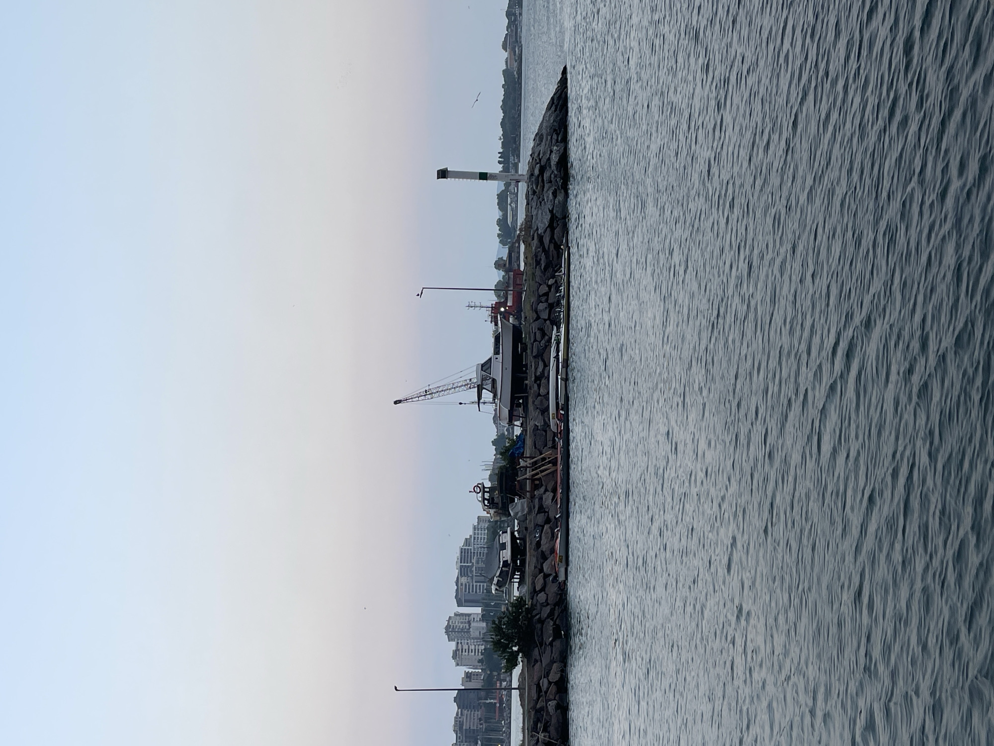 The image shows a harbor with several boats on the shore, as well as a lighthouse and a crane. In the background, there are buildings that appear to be part of the Kadıköy district in Istanbul, Turkey. The water is calm, and the sky is a soft blue, suggesting it is either early morning or late evening. The scene appears to be a working harbor, with boats of various sizes and states of repair. The overall impression is one of a tranquil, slightly industrial waterfront.