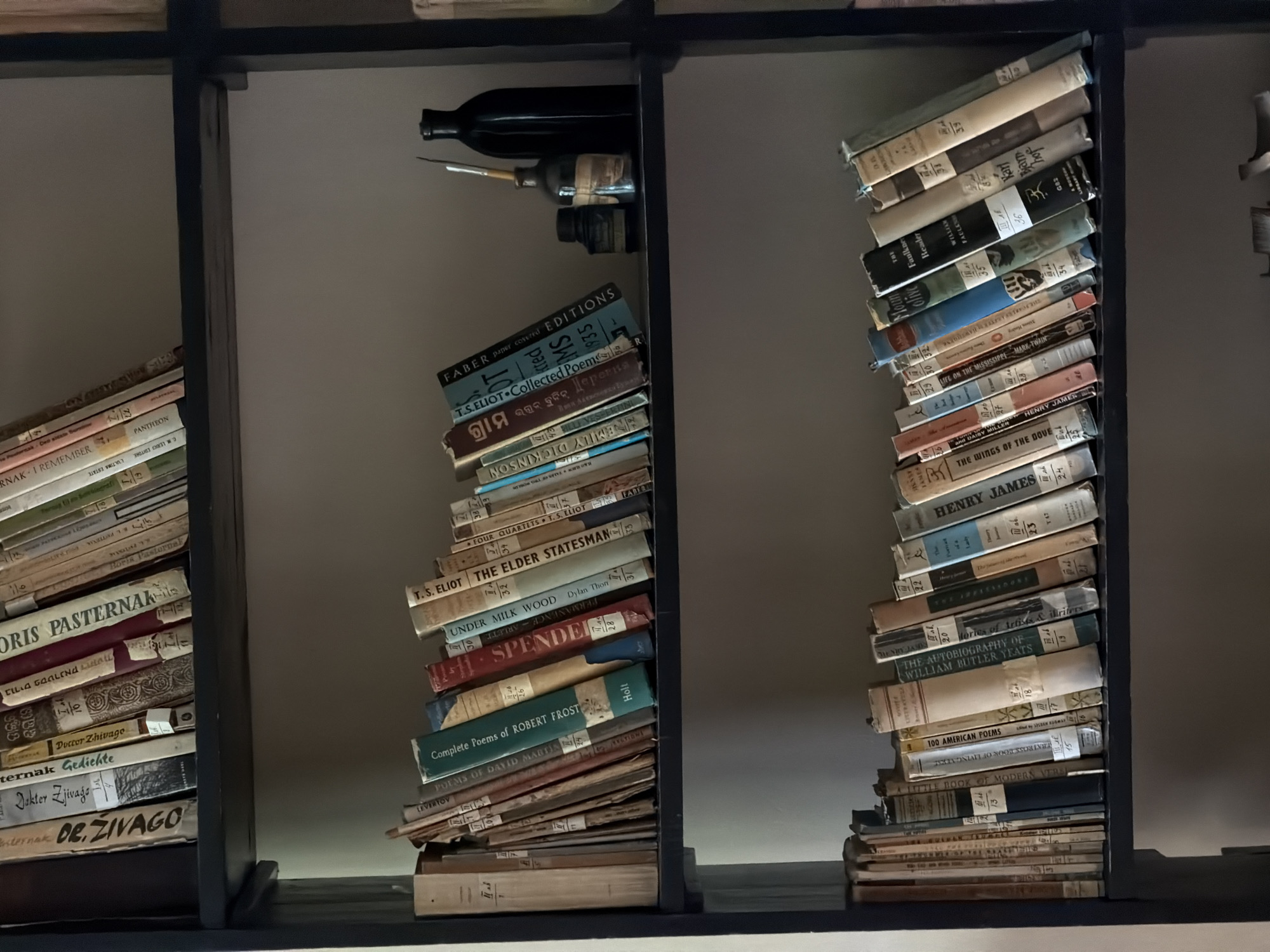 The image shows a dark wooden bookshelf containing three compartments. The left and right compartments contain stacks of books leaning precariously. The middle compartment contains stacks of books leaning precariously, and above them are three dark-colored bottles and a painting brush. The books have various titles, including works by Boris Pasternak, T.S. Eliot, Robert Frost, Henry James, and William Butler Yeats.
The location of Odintsovo, Russia, is not explicitly mentioned in the text of the books.