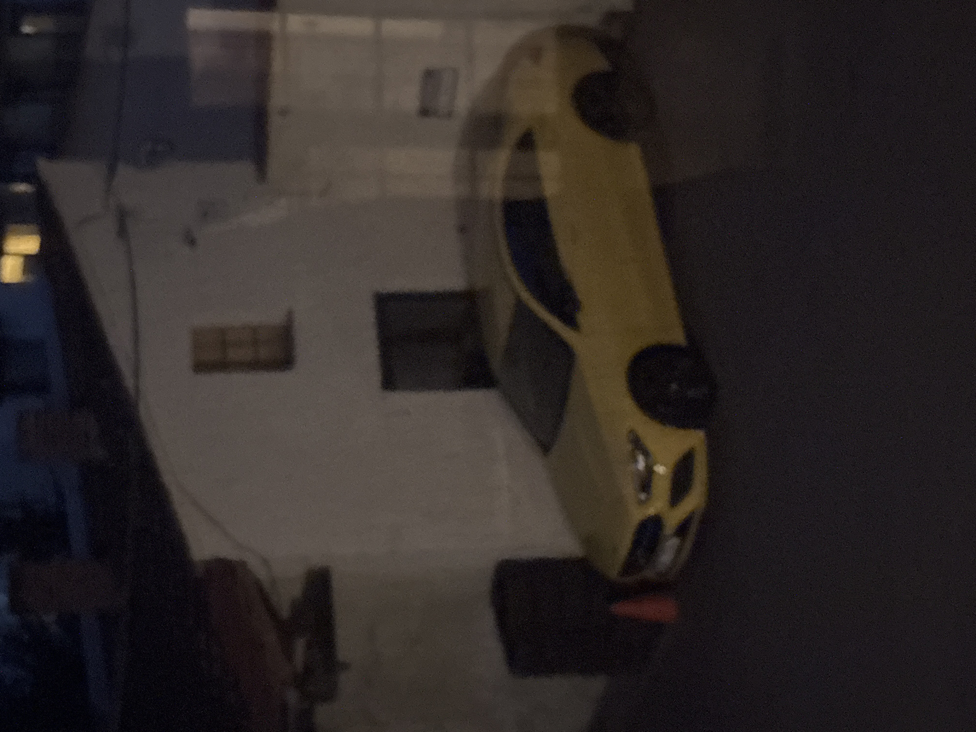 A yellow car is parked in front of a white building. The car is a coupe with dark tinted windows. A small orange traffic cone is visible to the left of the car. In the background, there is a dark building with lit windows. The scene appears to be outdoors at night.
