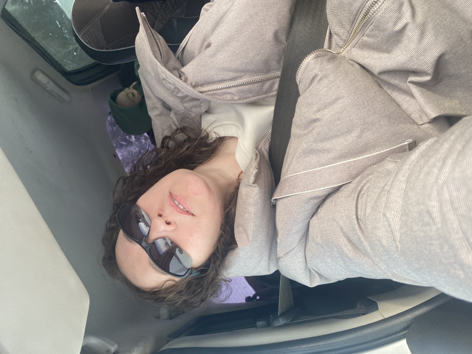 The scene is situated inside a vehicle, reportedly within the city of Ошья, Russia. An adult female is prominently featured in the foreground, occupying the front passenger seat, angled upward as if taking a selfie. She has dark, wavy hair and is wearing dark-tinted sports-style sunglasses, a light-colored crew-neck top, and a light-mauve or taupe-colored quilted winter jacket with a visible zipper closure. A dark-colored seatbelt diagonally crosses her torso. In the background, behind the woman and to her right, a child is partially visible in a dark-colored car seat, wearing a dark green hooded garment. The vehicle's interior ceiling is light-colored. Through the rear window, blurred outdoor elements, consistent with vegetation under daylight conditions, are discernible.