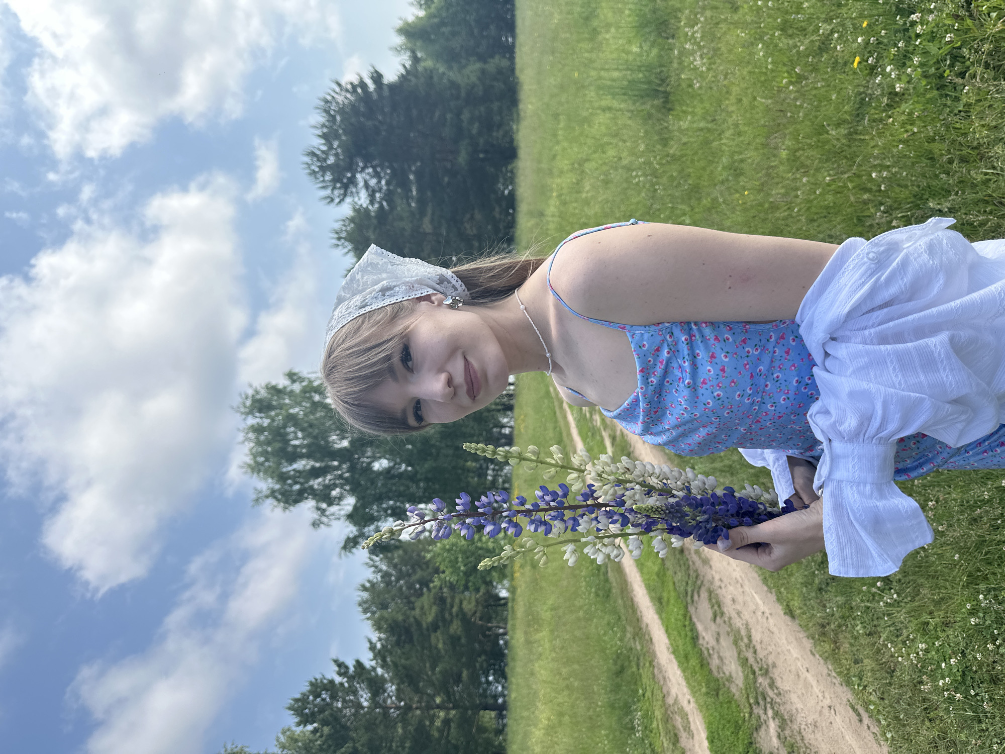 A young woman stands in a grassy field with a dirt path winding through it. She is wearing a blue dress with a floral print and a white headscarf. She is holding a bouquet of lupine flowers in her right hand. The background is a forest of pine trees under a cloudy sky. The woman is looking at the camera and smiling. The location is the city of Chaikovsky, Russia.
