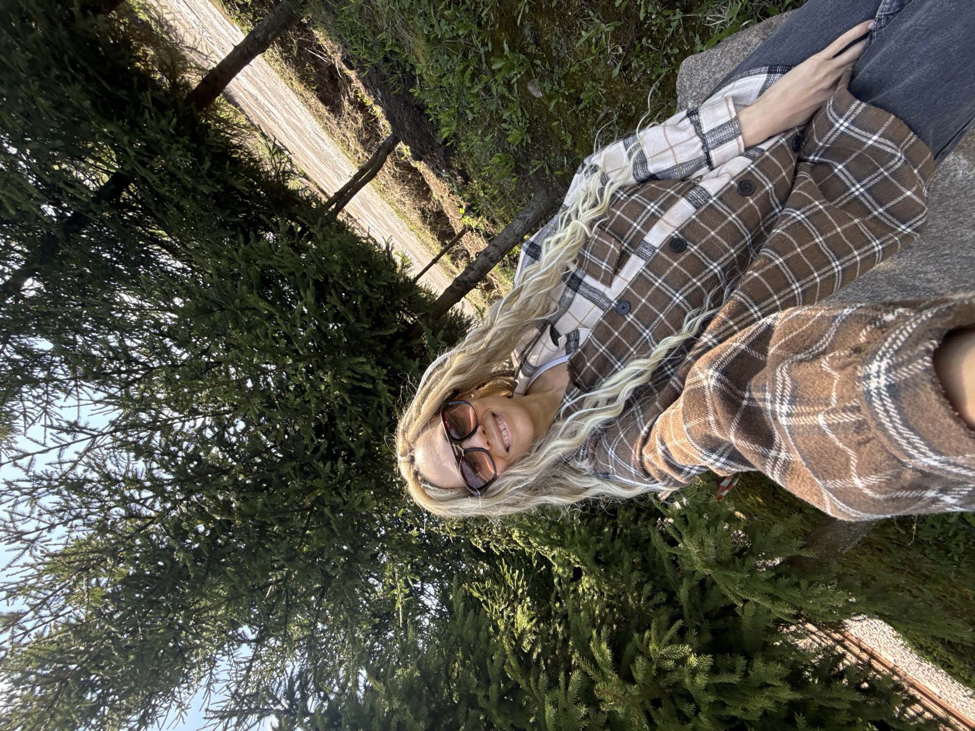 A woman with long, blonde dreadlocks is sitting outside near a large evergreen tree in Rusanovo, Russia. She is wearing sunglasses and a brown and white plaid shirt, and black jeans. She's posed for a selfie. A road can be seen in the background.