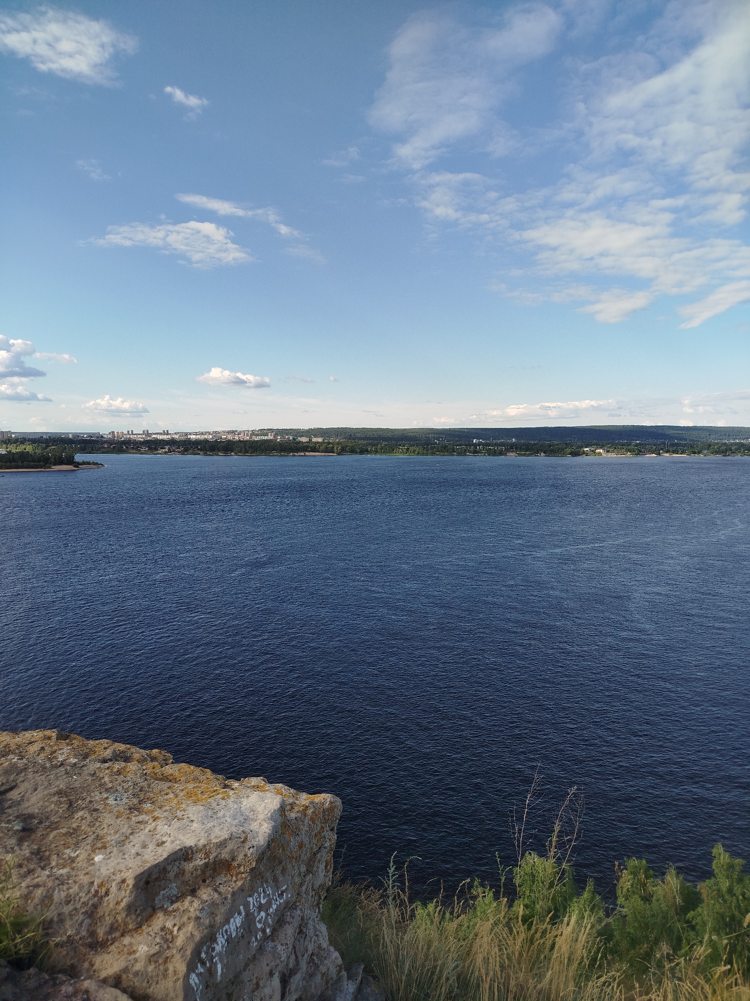 This media file captures a scenic view of a wide, deep blue river from a rocky outcrop. The water has a rippled texture, suggesting a gentle breeze. In the distance, across the river, is a densely forested shoreline that gives way to a cityscape with buildings visible against the horizon. The sky is a bright, clear blue with scattered white cumulus clouds. The foreground features a large, rough-hewn rock with patches of moss and lichen. Faint white graffiti is visible on the rock's surface. Dry grass and green bushes grow along the edge of the outcrop.
The scene depicts a tranquil landscape, likely during the daytime given the bright sunlight illuminating the sky and water. The presence of a city in the distance indicates a populated area, while the natural elements of the river, forest, and rock formation suggest an outdoor recreational or viewpoint location. The overall mood is serene and picturesque. The graffiti on the rock includes legible text in Cyrillic. The text on the rock, "акмола 2021" can be deciphered. Given the location context of Zhigulyovsk, Russia, this view is likely of the Volga River.