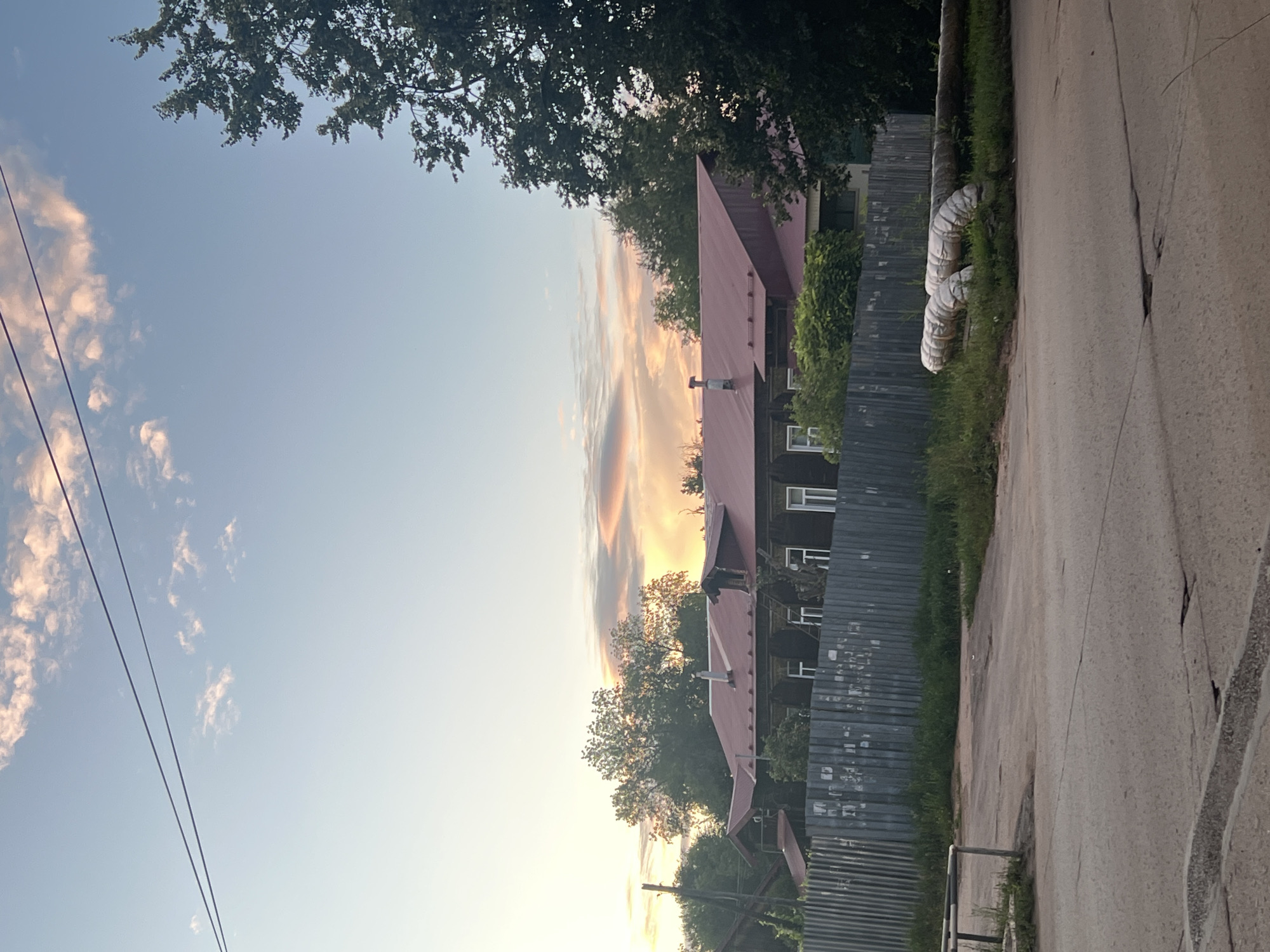 The image depicts a street scene in Ulyanovsk, Russia, at what appears to be dusk. A long, low-lying building with a red metal roof is visible behind a corrugated metal fence. The building has multiple windows and appears to be an older wooden structure. Trees with lush green foliage surround the building and extend into the background.
In the foreground, a paved road shows signs of wear and tear with cracks and patches. To the right of the road, large, insulated pipes are laid on the ground, snaking through the grass. A metal barrier with reflective strips is also visible on the left side of the road.
The sky above is a soft blue with wispy clouds, some tinged with pink and orange from the setting sun. Two dark power lines stretch across the upper portion of the frame. No people are visible in the scene. The overall impression is one of a quiet, possibly rural or suburban, street in the late evening.