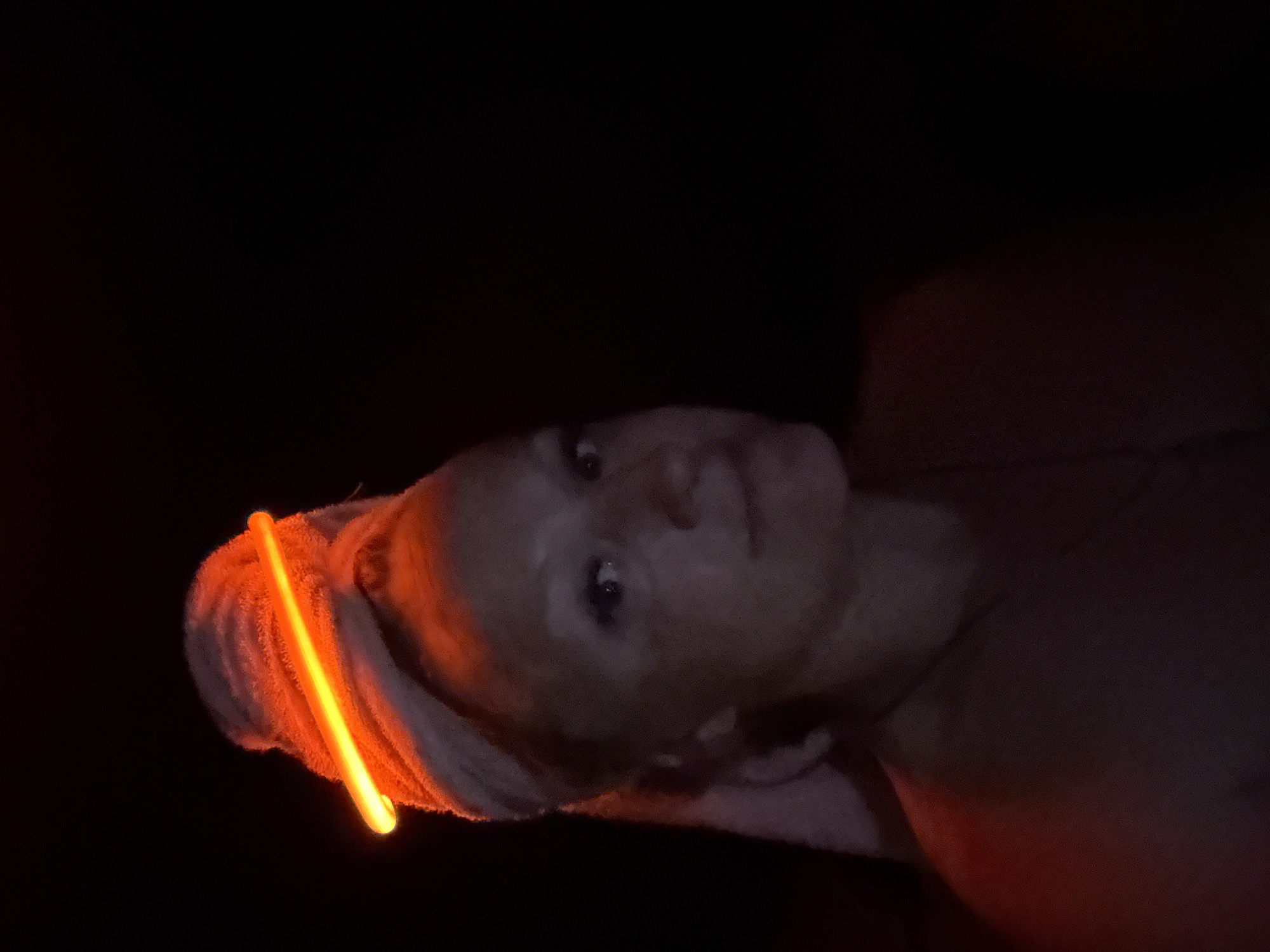 The image is a portrait of a person, oriented sideways, with their head positioned on the left side of the frame. The scene is exceptionally dark, with the background being almost entirely black, indicating an indoor setting at night or in a very dimly lit room.
The primary light source is a bright orange glow stick, which is tucked into a light-colored towel wrapped around the person's head. The glow stick casts an intense orange light onto the towel and illuminates parts of the person's face. The person's face is visible, looking slightly upwards towards the right side of the frame, with a neutral and calm expression. Their eyes are dark and appear to be open. Due to the low light, specific details of their facial features or skin texture are indistinct, though their skin appears light-toned. Their shoulders and upper chest are also dimly lit by a soft, warm orange glow.
No explicit activity is depicted other than the person relaxing or engaging in a self-care routine, possibly post-shower or with a face mask, given the towel and intimate setting. There are no discernible location cues beyond the dark indoor environment. No text is visible in the image. The overall mood conveyed is one of quiet relaxation or introspection, highlighted by the soft, singular light source.