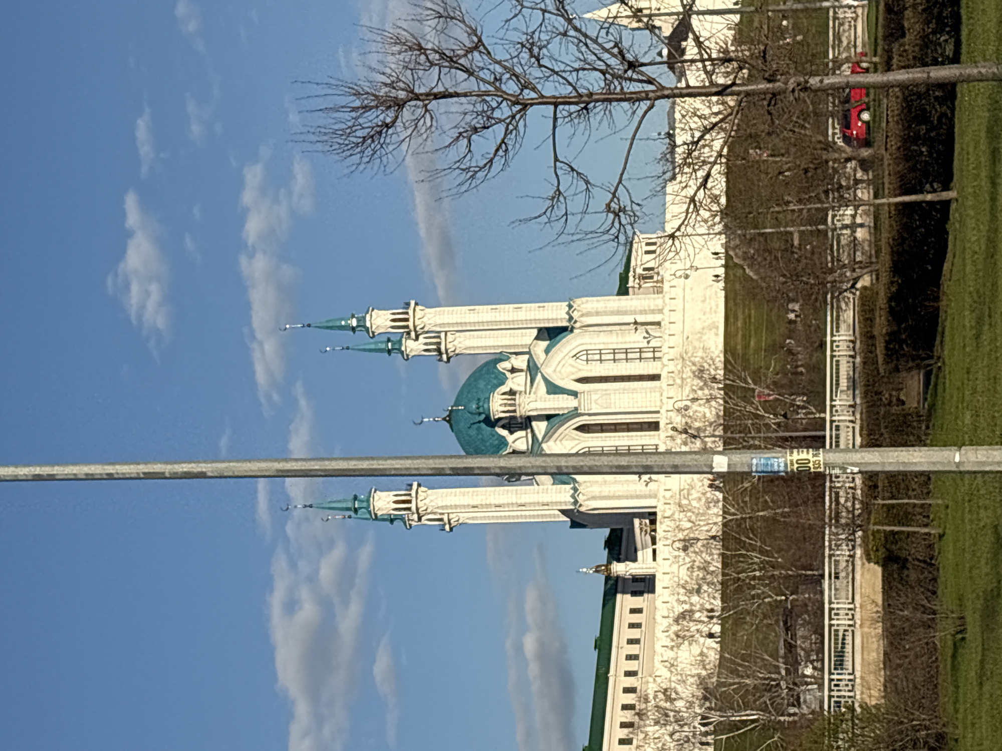 The image depicts the Qolsharif Mosque, situated in the city of Kazan, Russia. The mosque is constructed in white stone with a turquoise dome and spires. The sky is light blue with some clouds. A bare tree is visible in the foreground and a street lamp in the lower left. The mosque is positioned near a green lawn or park area. A red car is visible near the building and there are some people walking in the vicinity.