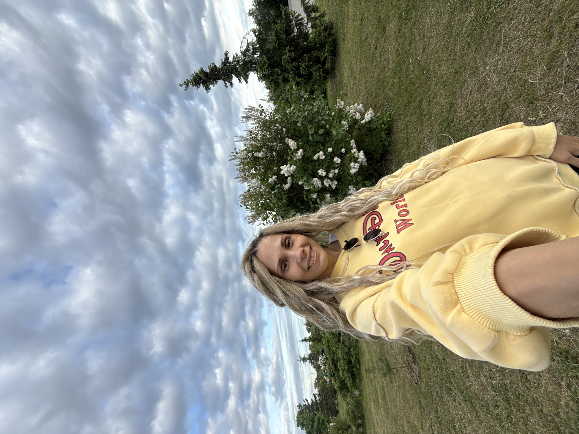 A woman is featured in a selfie, smiling directly at the camera. She has long, wavy blonde hair, with some sections styled into dreadlocks or thick braids. She is wearing a bright yellow long-sleeved sweatshirt with red text, where the word "World" is clearly visible, and parts of other words can be seen. Sunglasses are hooked onto the collar of her sweatshirt.
The setting is outdoors, in what appears to be a park or a grassy public area in Шебунино, Russia. The background includes green grass, several trees (including conifers), and a bush with small white flowers. In the distance, behind the trees, a large Ferris wheel-like structure is faintly visible. The sky is largely covered with grey and white clouds, but patches of blue sky can be seen peeking through, indicating it is daytime, possibly with overcast weather. The woman's joyful expression suggests a positive mood.