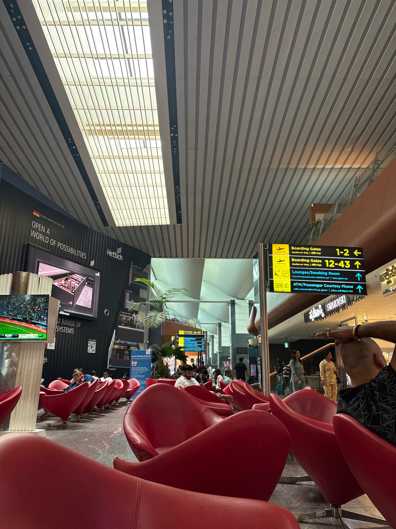 The image depicts the interior of an airport waiting area. The dominant feature is a row of bright red, modern-style chairs arranged in a curve. Sunlight streams in through a large, slatted skylight system in the ceiling. On the left, a large screen displays what appears to be a cricket match. Behind the chairs, a black wall features branding for "Hettich" and text that reads "OPEN A WORLD OF POSSIBILITIES". To the right of the seating area, a directional signpost provides information in multiple languages, indicating boarding gates, lounges, and ATMs. Several people are seated in the chairs, some looking at their phones or conversing, while others are in the background walking or standing. The floor is a polished, grey stone. The overall atmosphere is one of a busy, modern airport terminal.