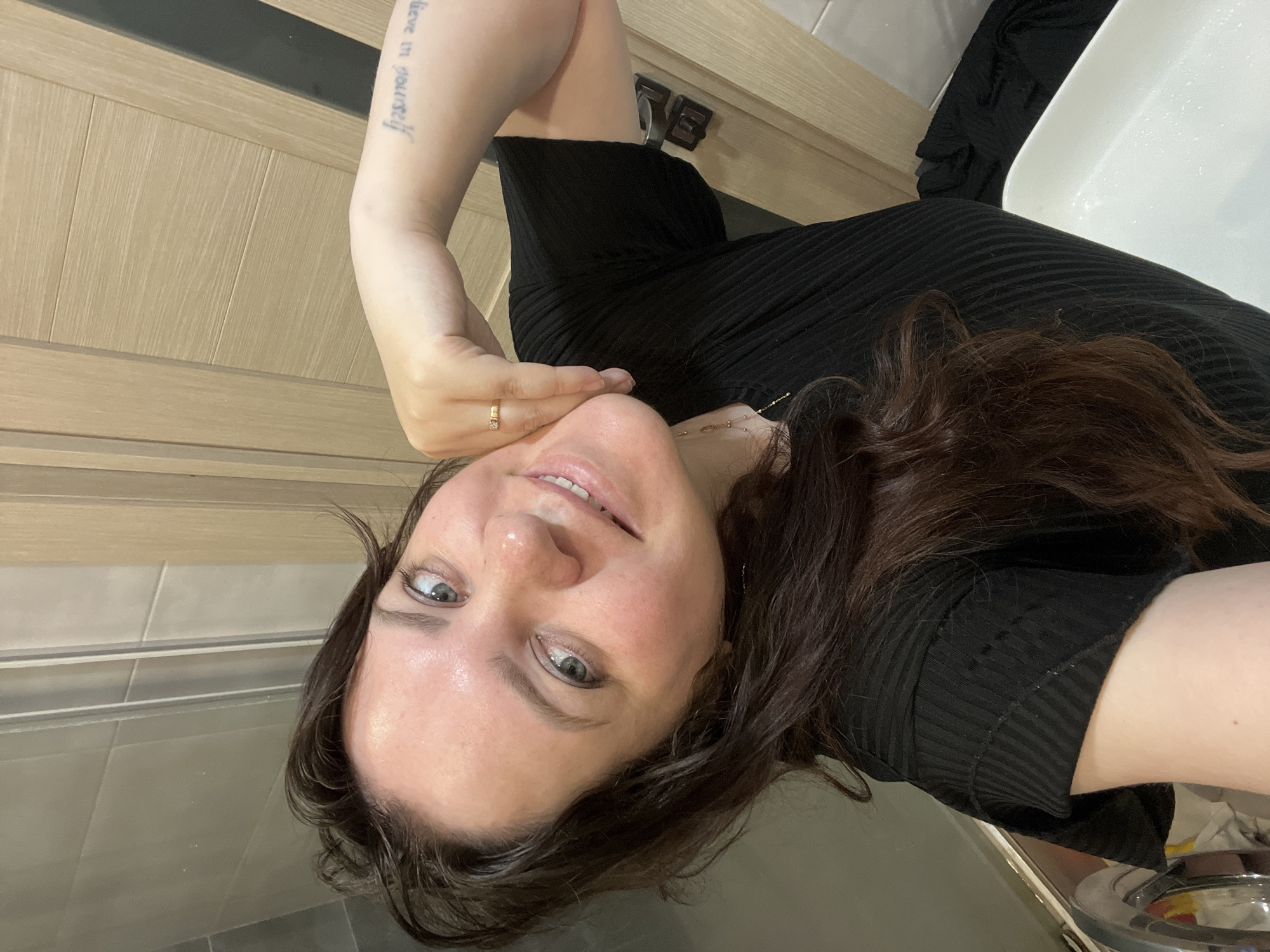 A selfie was taken in what appears to be a bathroom with white ceramic tiles and a wooden door. A woman with fair skin, shoulder-length brown hair, and grey eyes is posing. She is wearing a black ribbed top. Her left arm is raised, and a gold ring is visible on her ring finger. A tattoo is on her forearm, but the text is difficult to decipher. In the background, a white sink and parts of bathroom fixtures are visible. The scene does not provide enough information to pinpoint a specific location in Noviy, Russia.