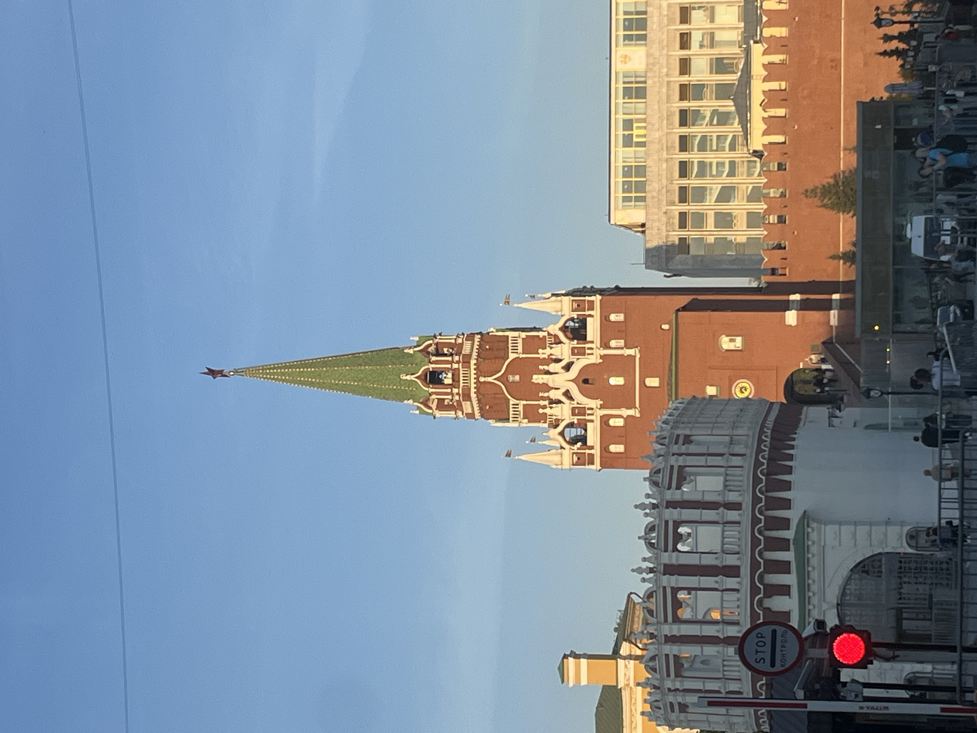 Here is a description of the image.
The image shows a view of the Moscow Kremlin, a fortified complex at the heart of Moscow, Russia. Visible is one of the Kremlin's towers with a distinctive green roof and a red star on top. The tower is made of red brick and adorned with white decorative elements. Other buildings within the Kremlin are visible, also made of red brick with white details.
A lighter-colored building stands behind the Kremlin structures. In the foreground, there is a white structure with red decorative stripes, appearing to be part of the Kremlin wall. A traffic barrier is present, along with a "STOP" sign written in Russian, and a lit red traffic light. This suggests an entrance point with security control. The sky is clear and blue, indicating a bright day.
