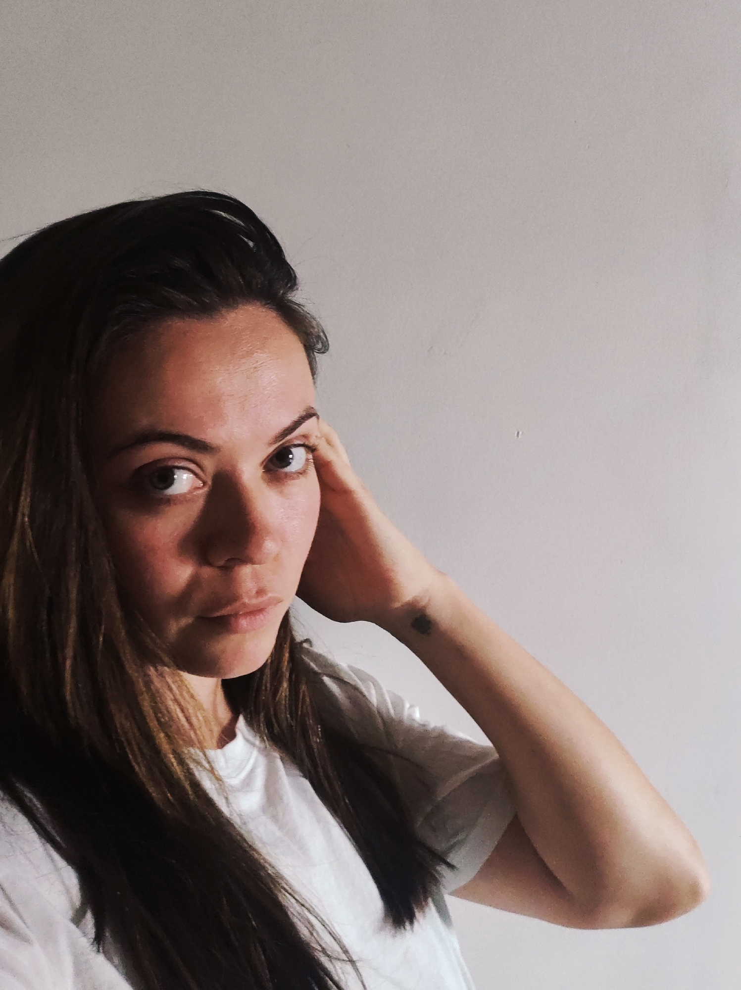 A medium close-up portrait features a young woman with long, dark brown hair, looking directly at the camera. She wears a plain white t-shirt. Her head is tilted slightly, and her left hand is raised, with her fingers gently resting near her temple/ear. A small, dark, possibly heart-shaped or irregular blob tattoo is visible on her left wrist.
Her expression is serious and direct, with a contemplative gaze. Her lips are slightly parted. The lighting appears soft, coming from the left, casting subtle shadows on the right side of her face and hair, creating some contrast.
The background is a plain, light-colored (off-white or light gray) wall, suggesting an indoor setting. There are no other objects or discernible features in the background. No text is visible in the image.