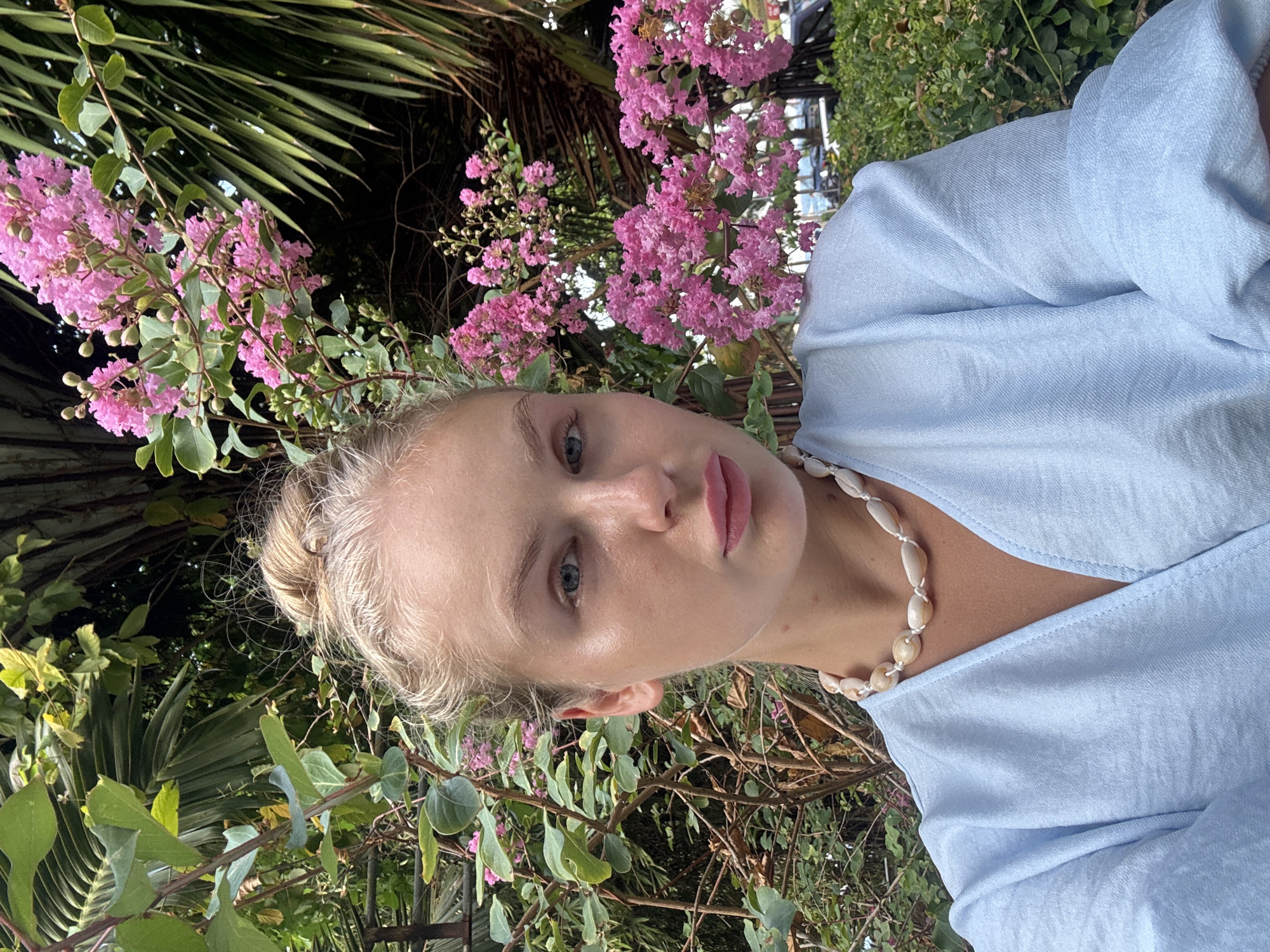 A medium shot shows a young blonde woman with her hair in a bun, wearing a light blue wrap top and a necklace made of shells. She is looking at the camera with a neutral expression, and her face is partially obscured by pink flowers and green foliage. The background is lush and green, with palm fronds visible. It is daytime and the lighting suggests an outdoor setting, possibly a garden or park. The woman is centered in the frame, with the flowers and foliage framing her face and upper body. The image appears to be a selfie.