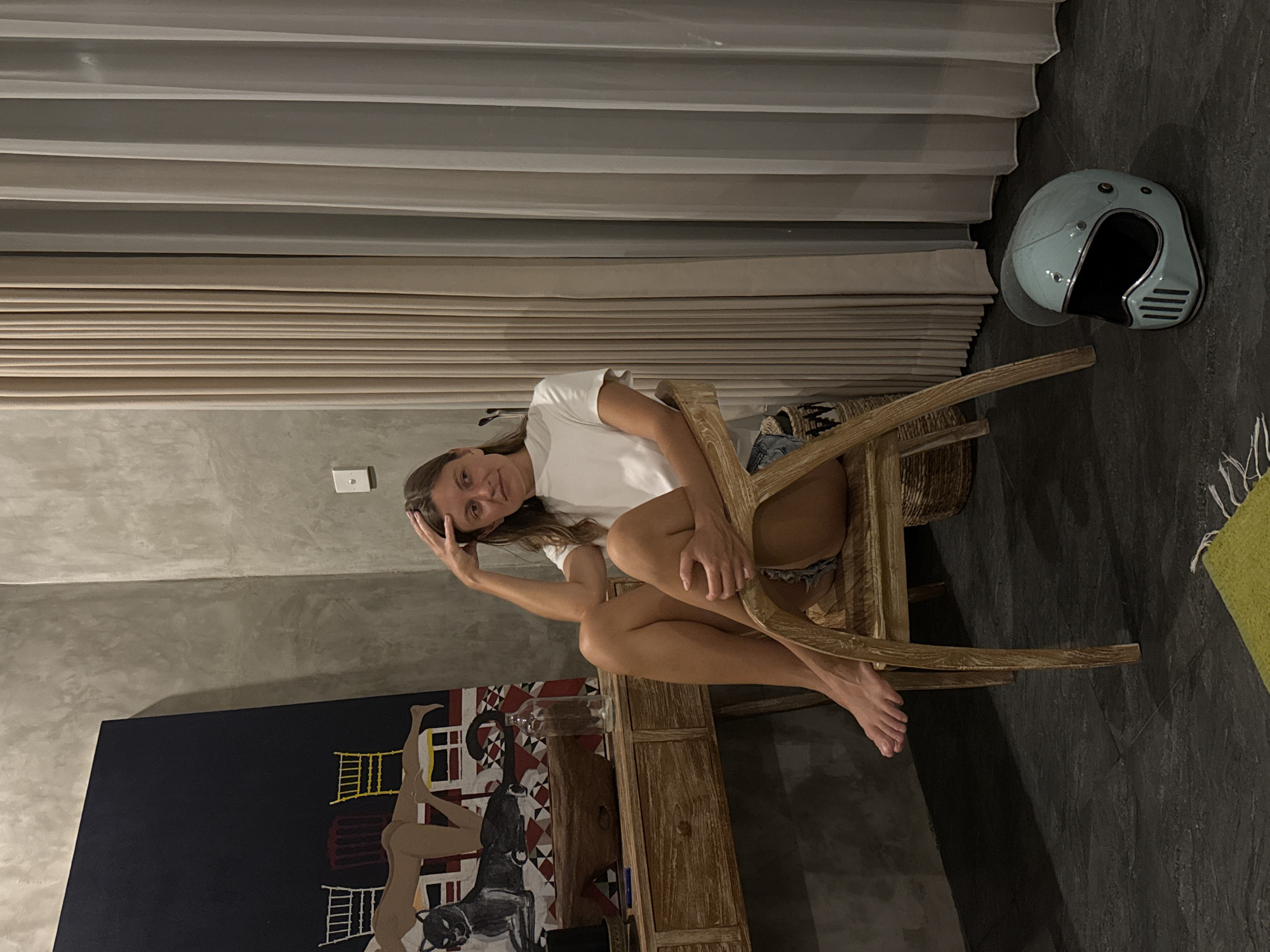 A woman sits in a wooden chair in an indoor setting. She wears a white t-shirt and denim shorts. She rests her head on her left hand. Behind her is a gray wall and beige curtains. To her right, a light blue motorcycle helmet rests on the floor. To her left, there is a painting on a blue canvas, showing a figure. The location is possibly in North Kuta, Indonesia.