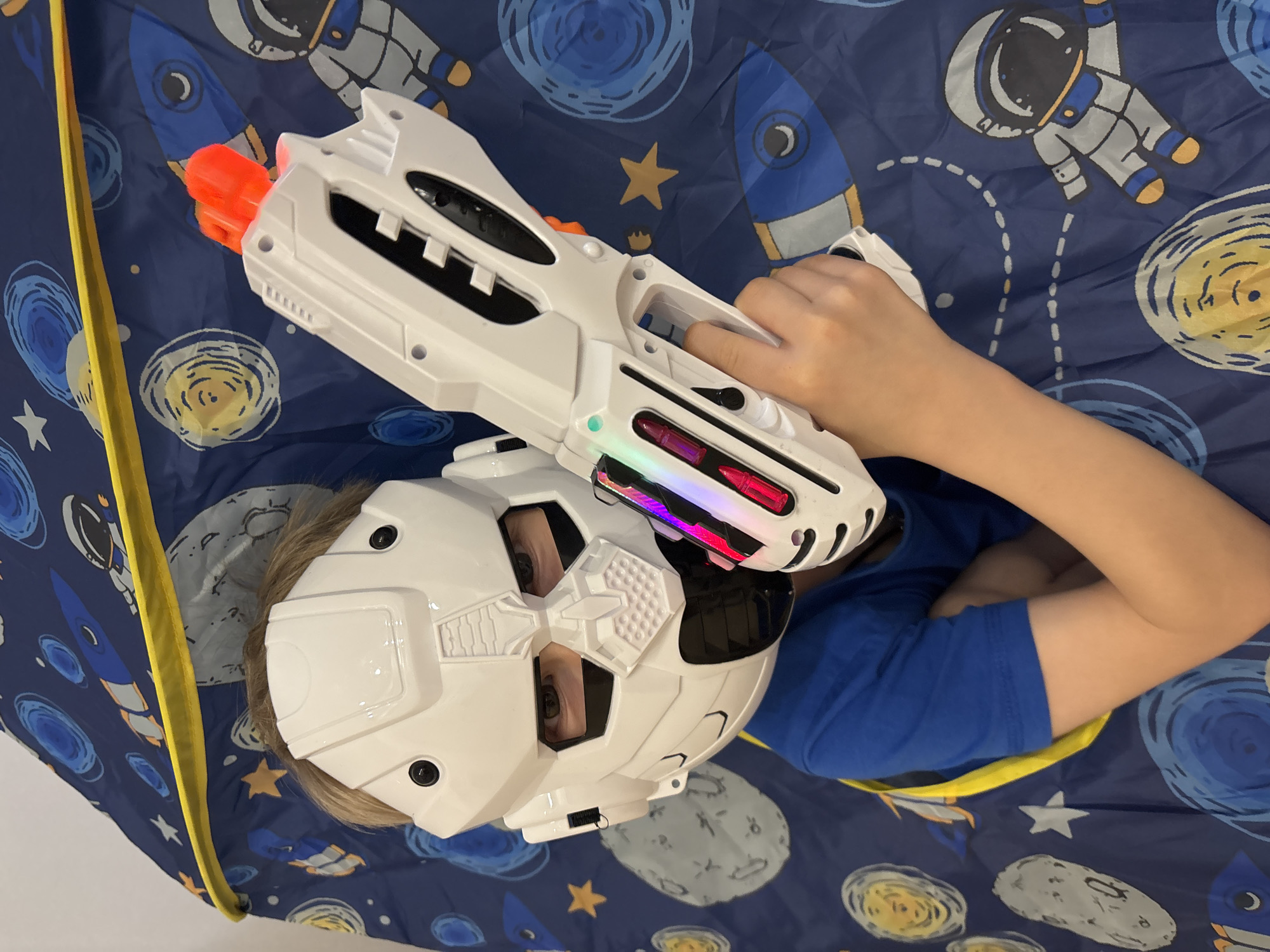 A child wearing a white helmet and holding a white toy gun with an orange tip lies inside a blue tent decorated with space-themed designs. The child is wearing a blue shirt. The location is implied to be Chaikovsky, Russia.